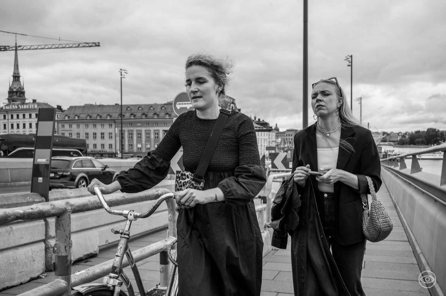 Two women walking on a bridge, one with a bicycle, black and white urban street photography.