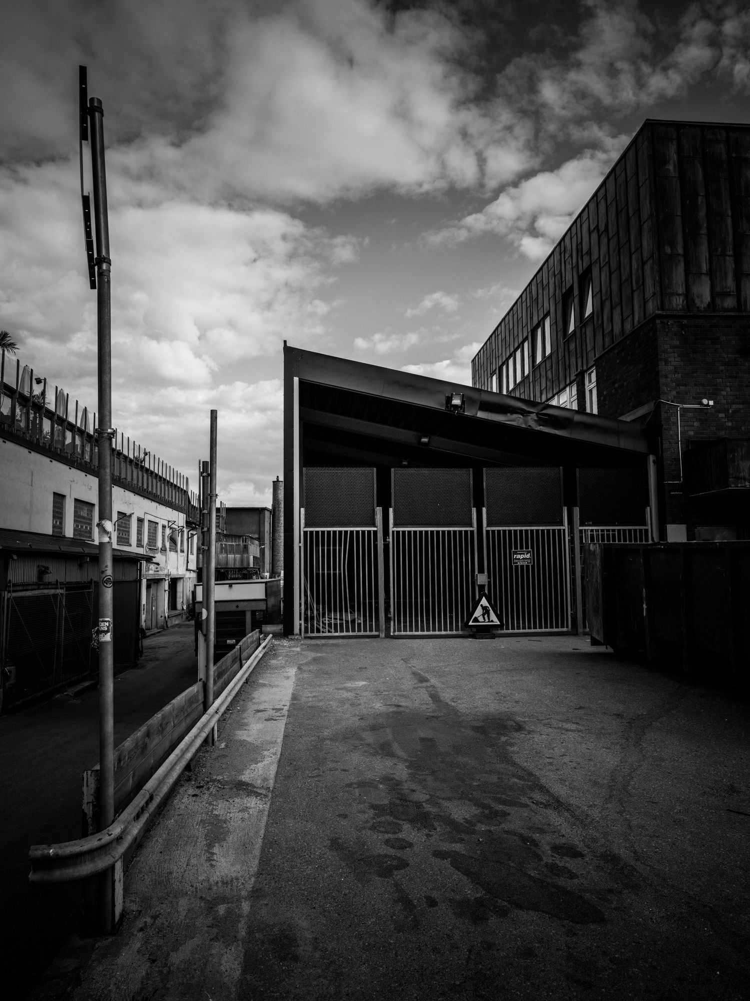 Black and white urban photograph of an industrial gated entrance with dramatic cloudy sky.