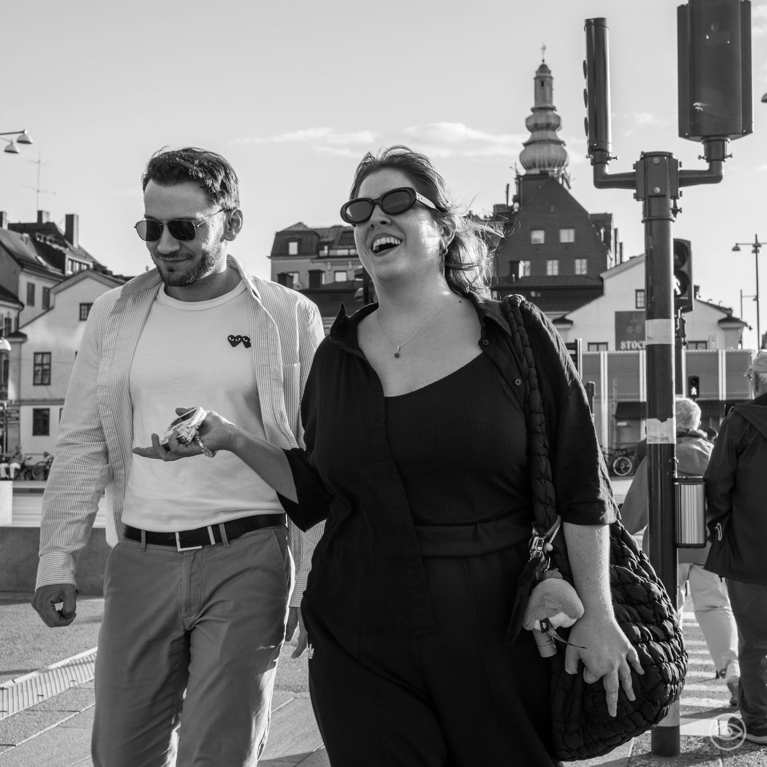 Laughing woman in sunglasses and man in white jacket walking together on a sunny street.