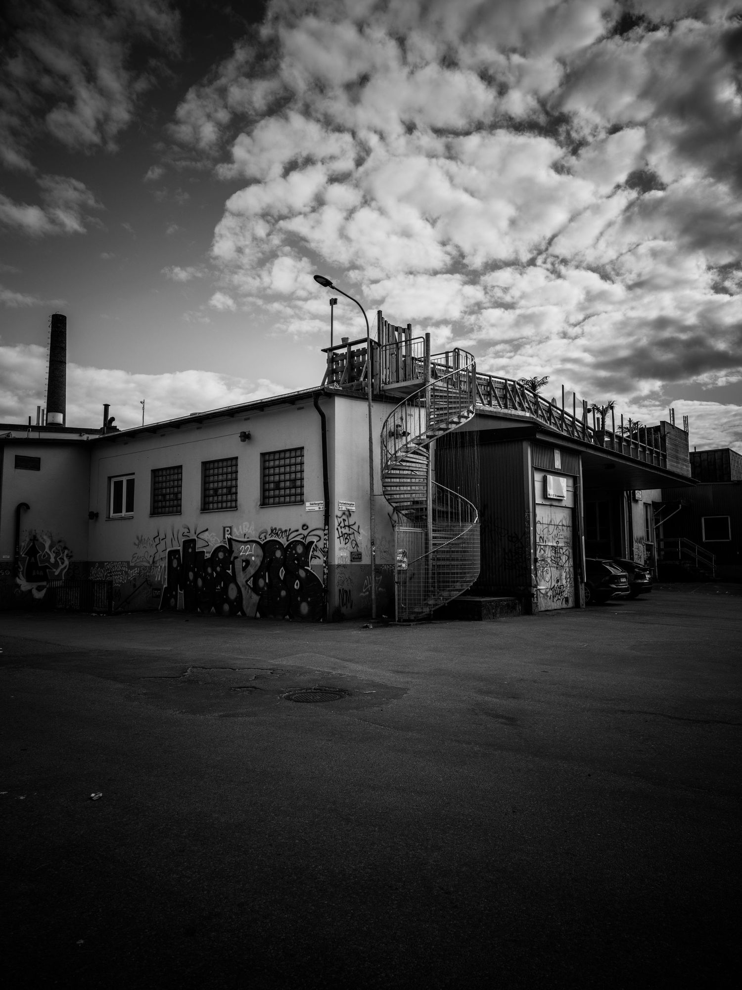 Black and white wide shot of an industrial building with spiral staircase under dramatic clouds.