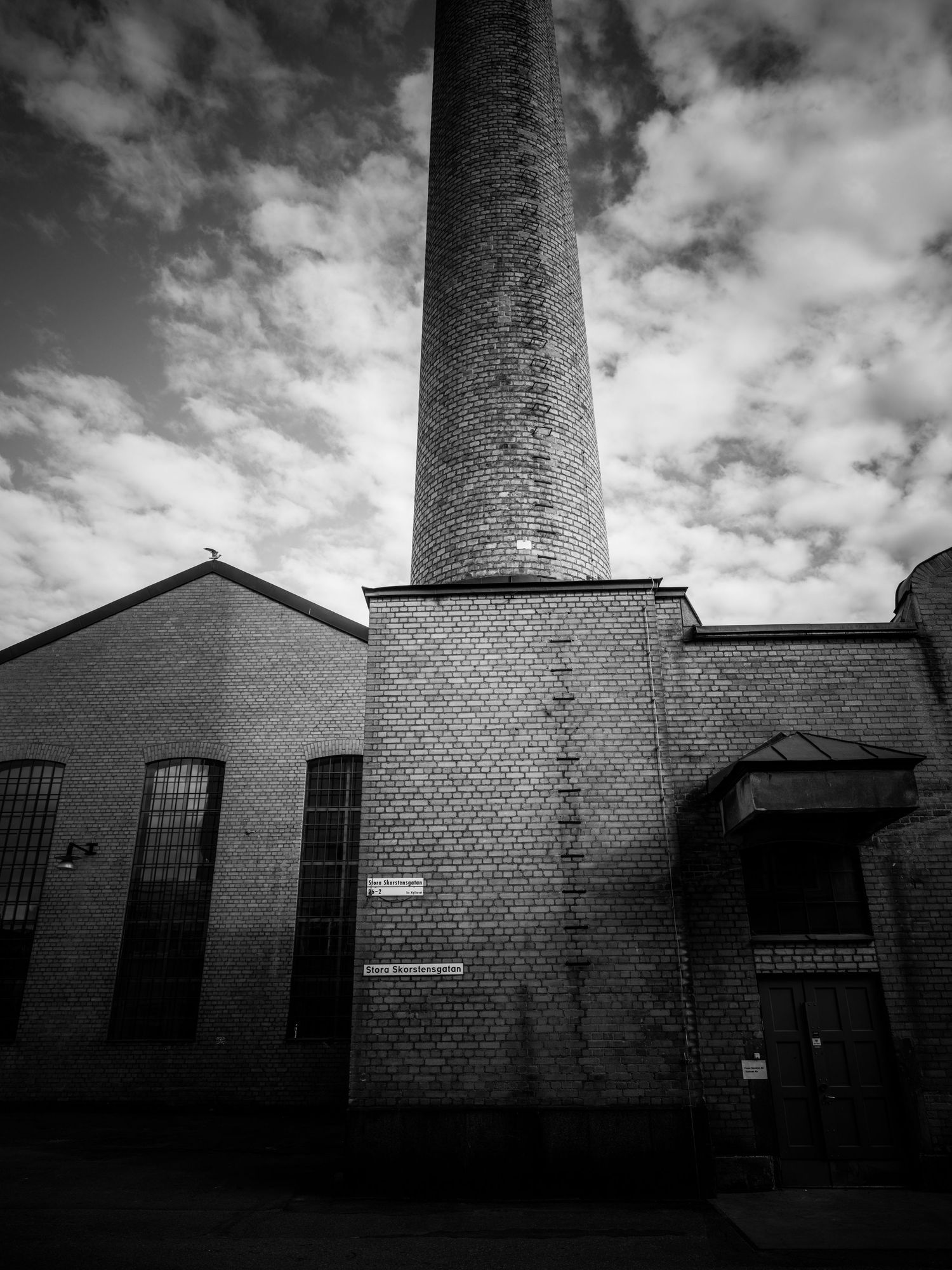 Tall brick chimney stack rising above an abandoned industrial building under dramatic sky.