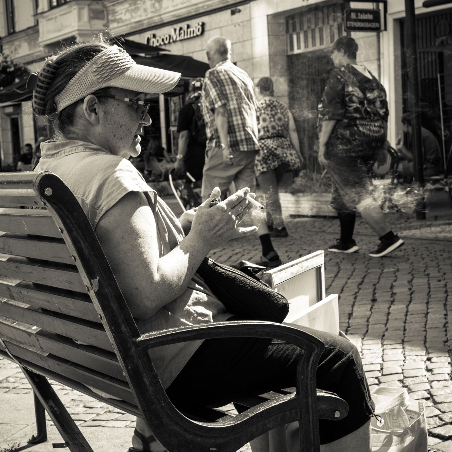 Woman in cap and sunglasses sits on bench eating in busy cobblestone street, black and white.