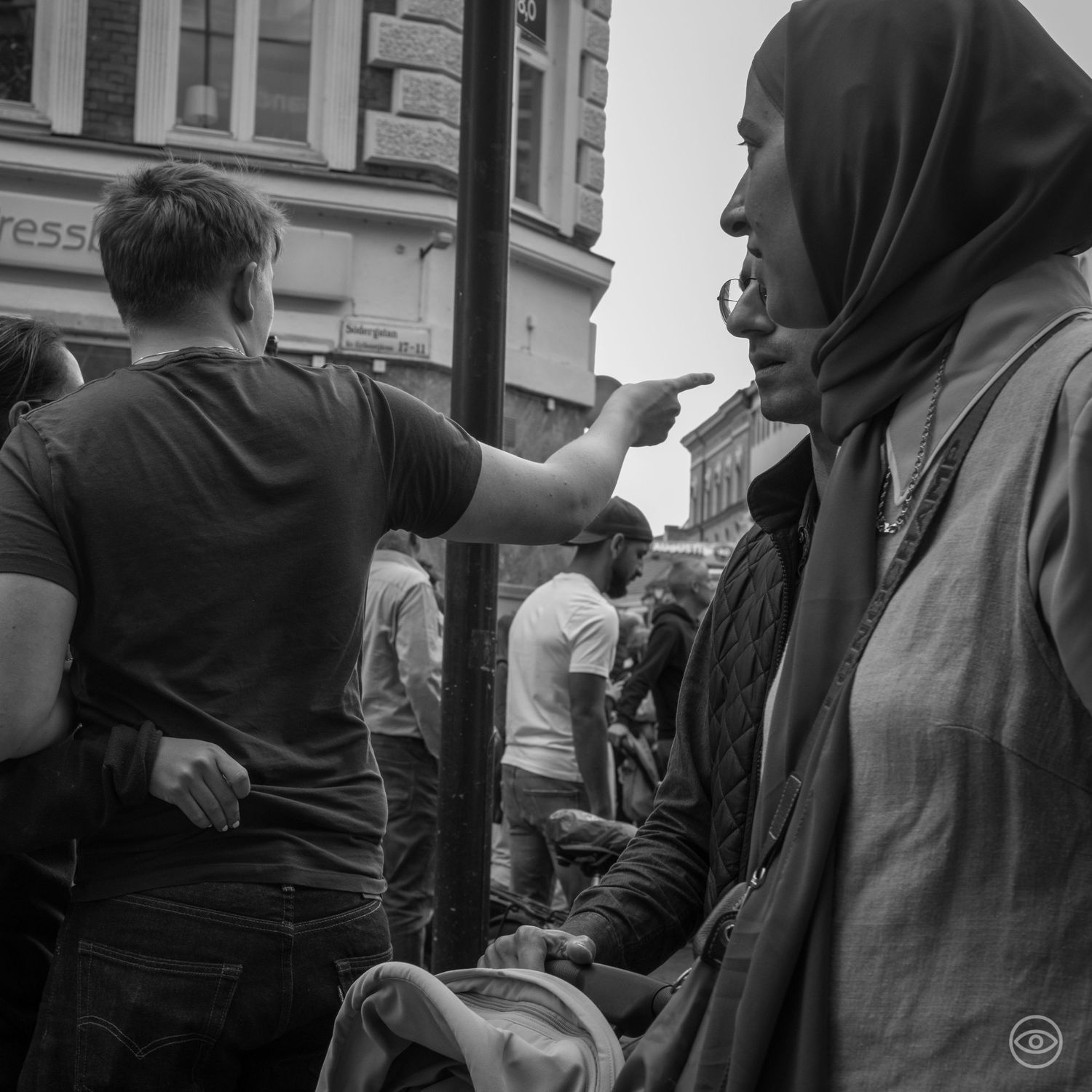 Black and white street photo of a crowd with a woman in hijab and a man pointing in an urban setting.