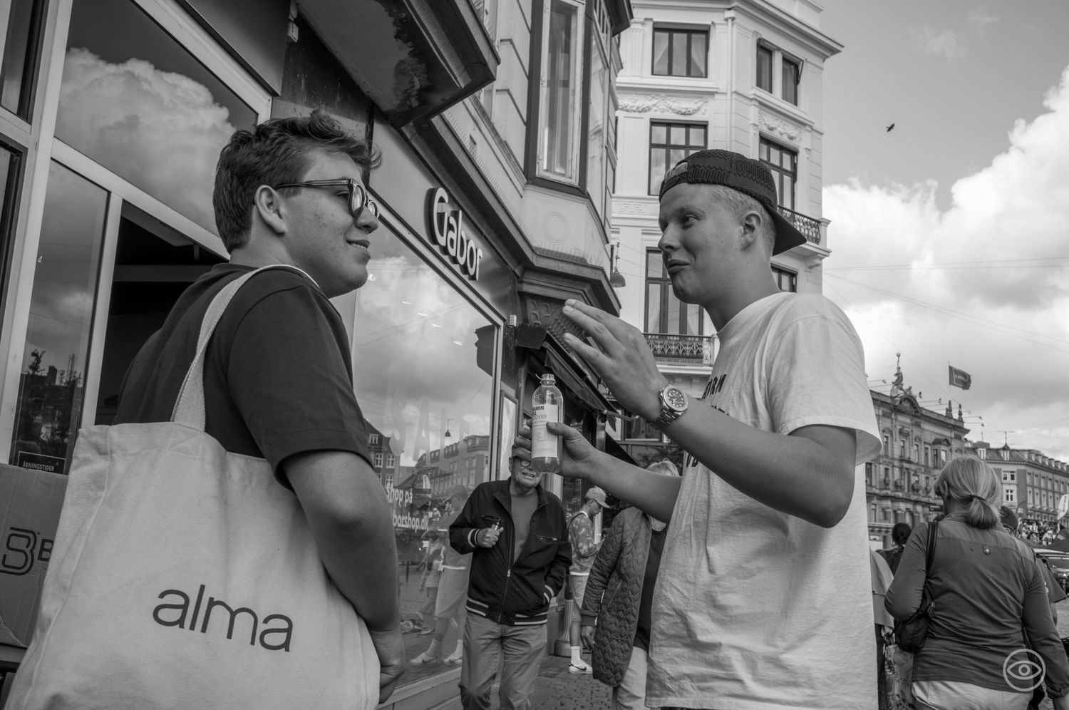Two men engaged in conversation on a busy urban street, one carrying a white Alma tote bag, black and white street photography.