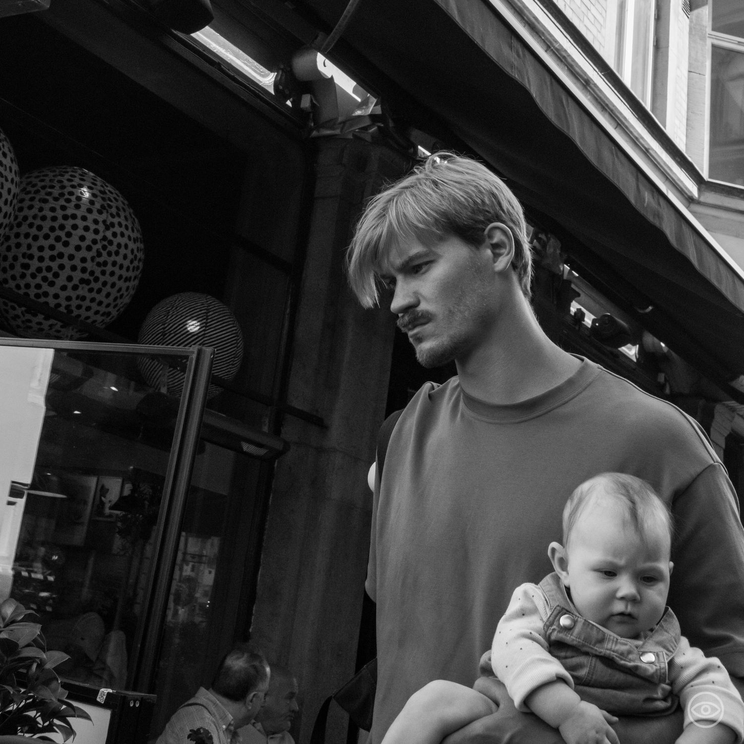 Young father holding a baby outside a shop entrance in a candid black and white street photo.