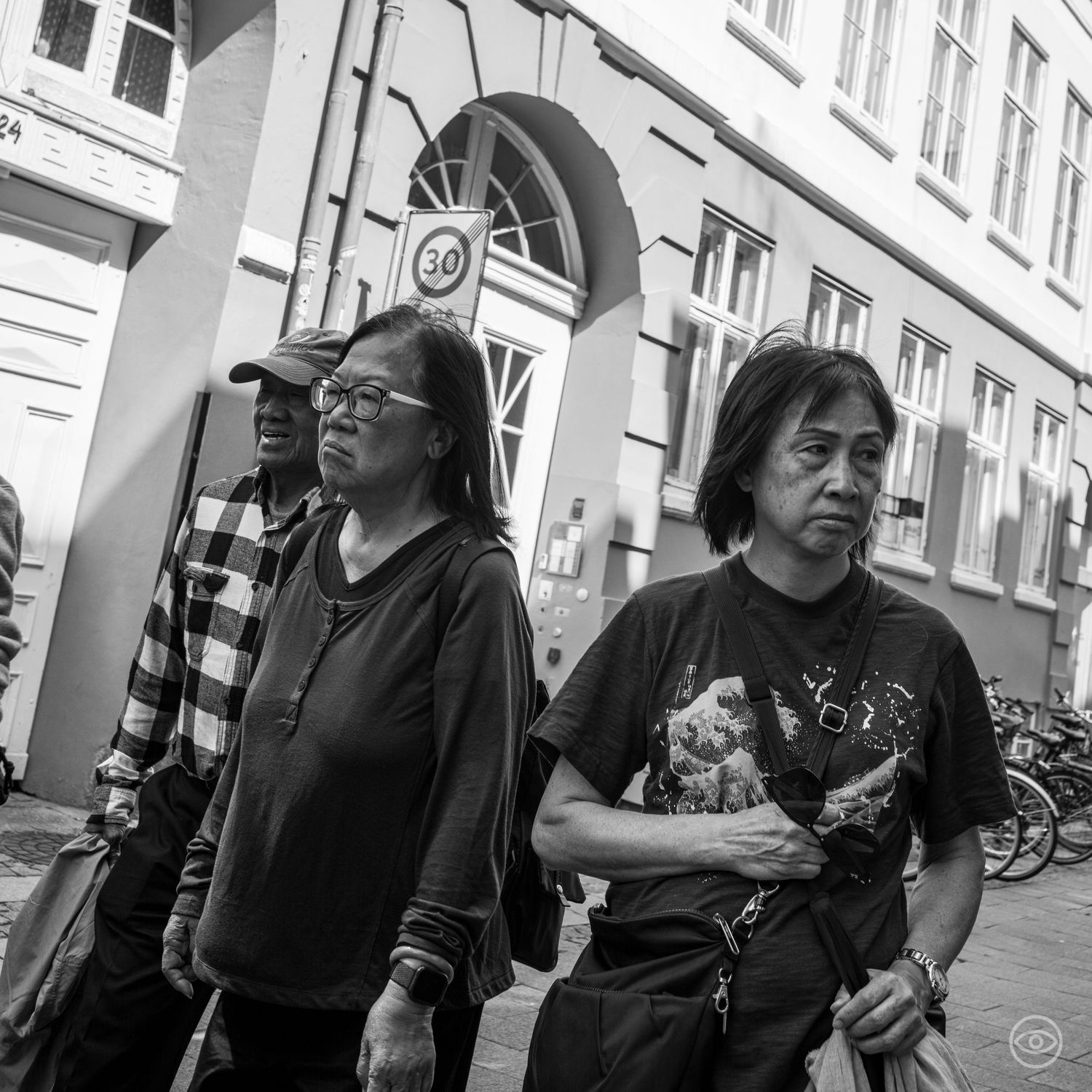 Three Asian pedestrians walking along a cobblestone European street past a 30 speed limit sign, black and white photography.