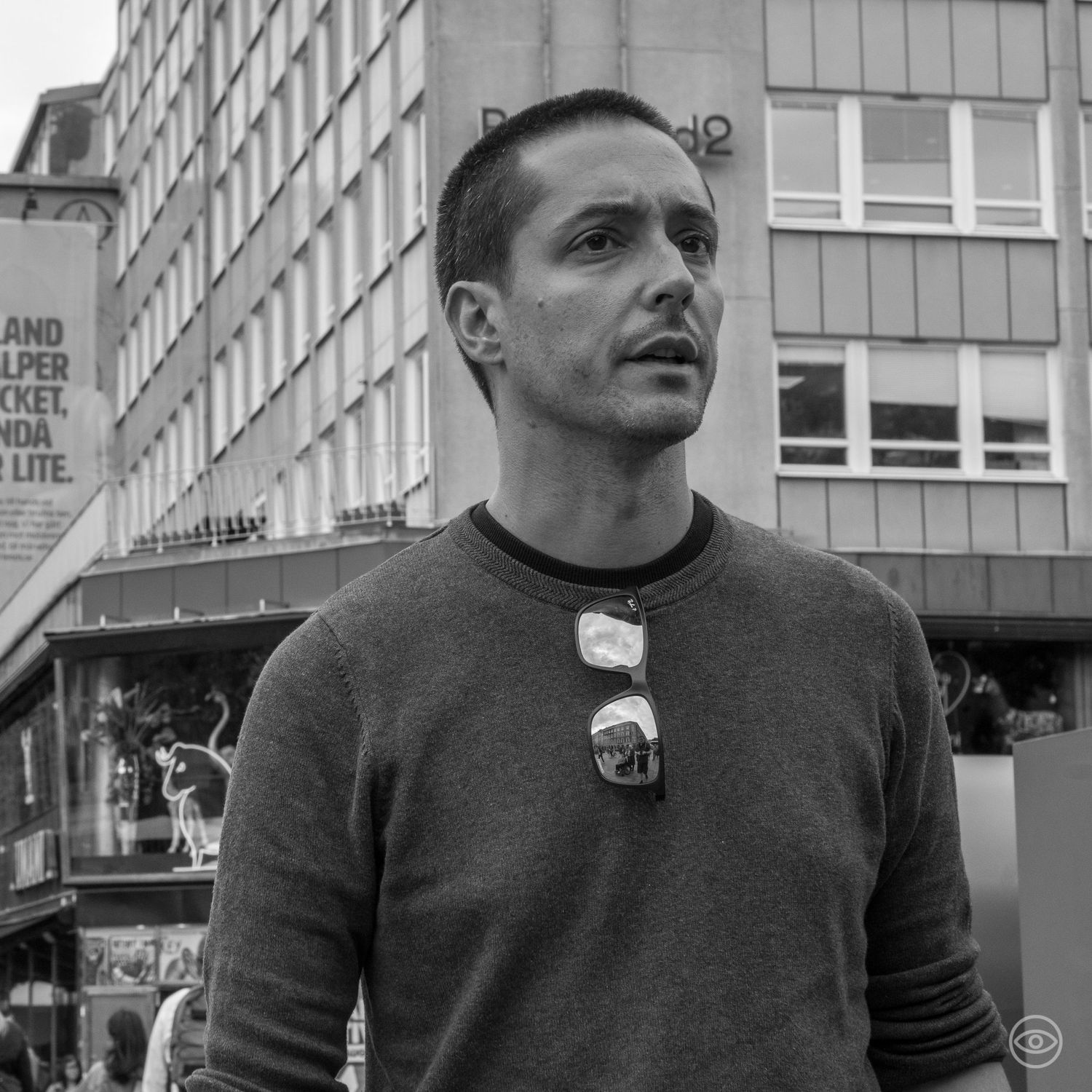 Man in a gray sweater with sunglasses hanging from collar standing in front of urban buildings.