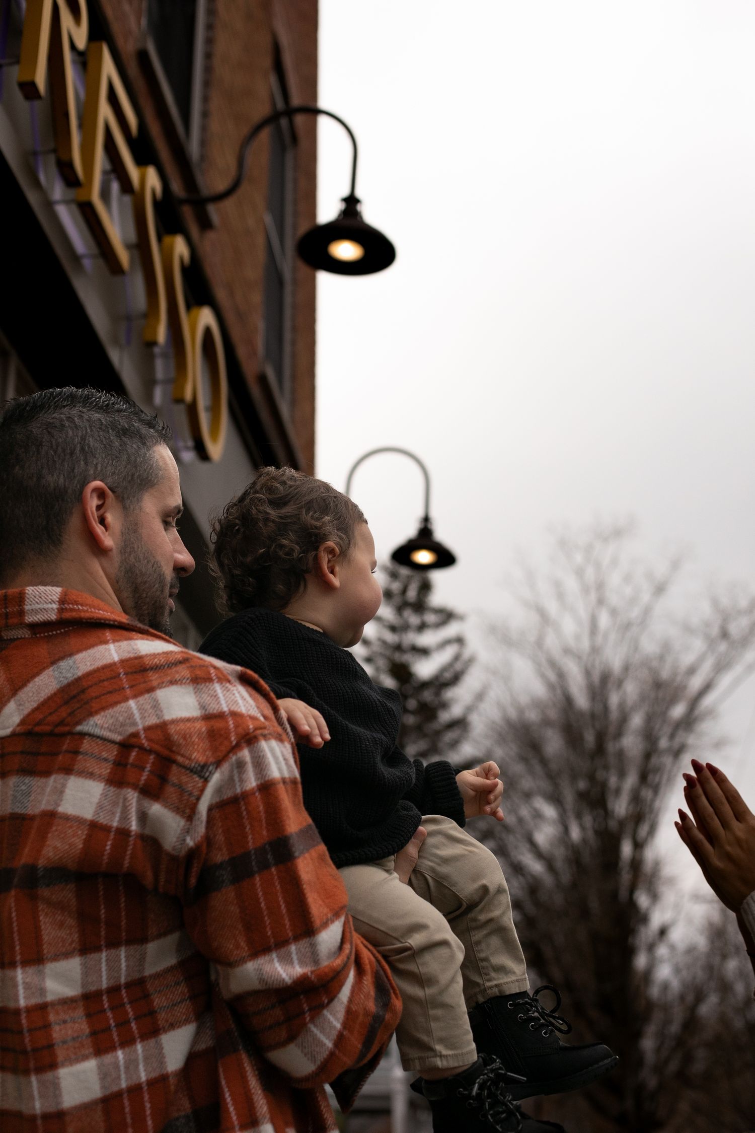 A father holds his young child outside a brick building with vintage street lamps on a cloudy day in Syracuse NY. By Candid House Studios.