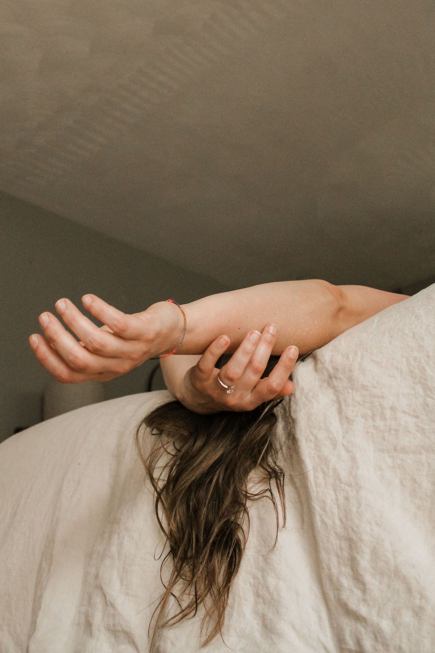 A woman with long hair hides under white linen bedding, her ringed hands reaching over the edge.