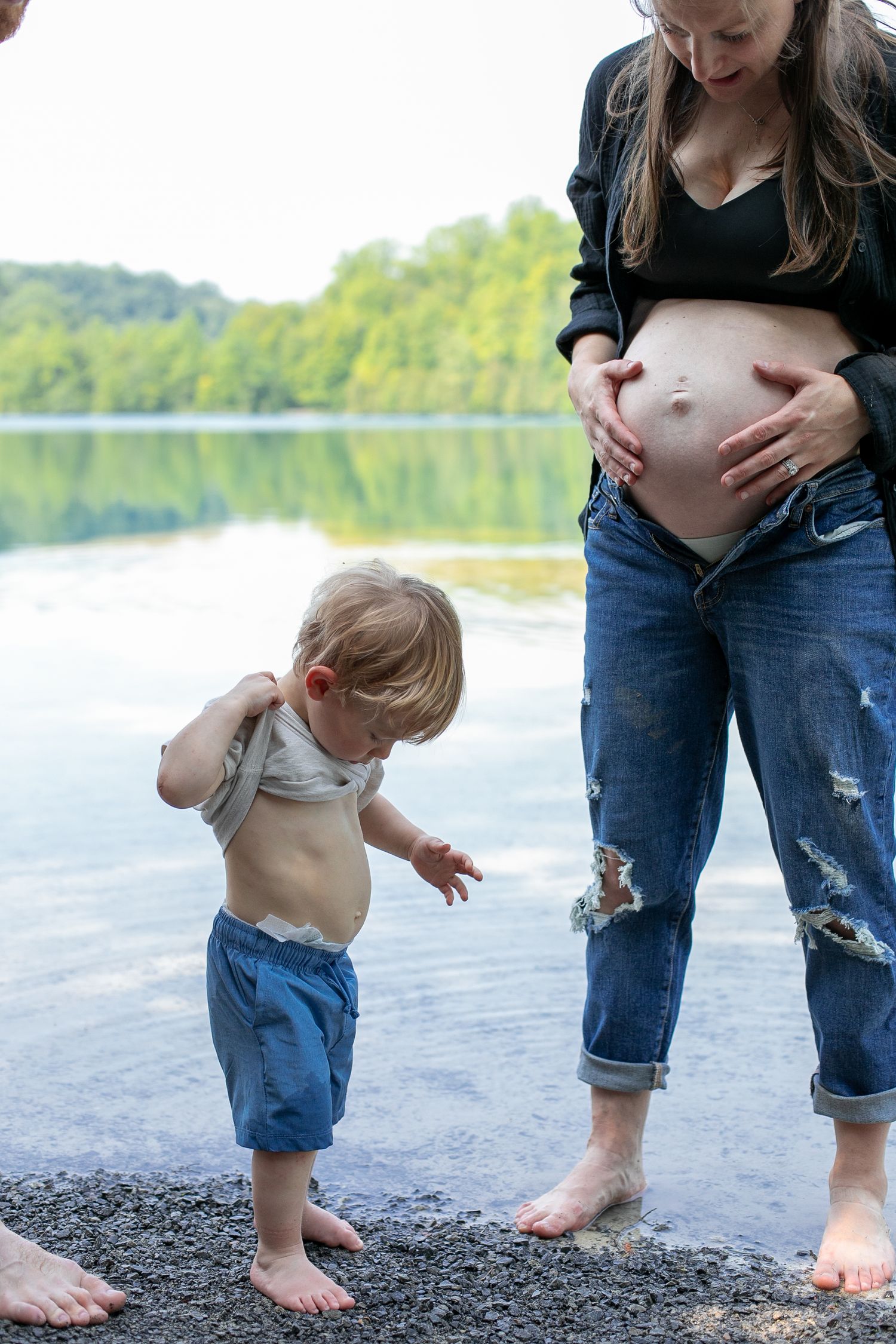 Pregnant mother in ripped jeans with toddler lifting shirt on rocky lakeside beach during maternity photos by Candid House Studios