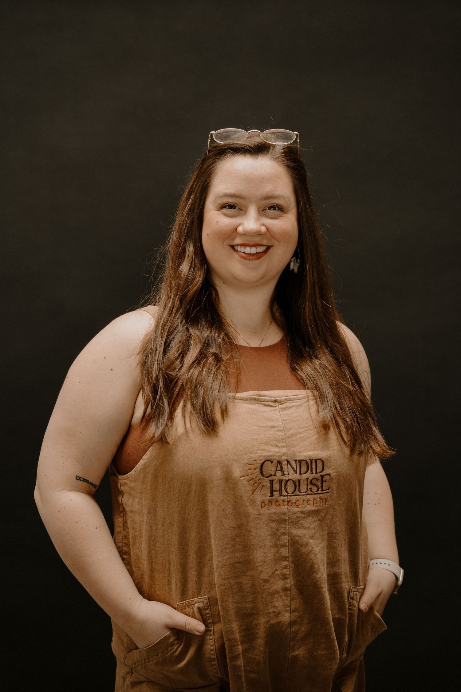Smiling woman with sunglasses on head wearing tan Candid House overalls, posing with hands in pockets against dark background.
