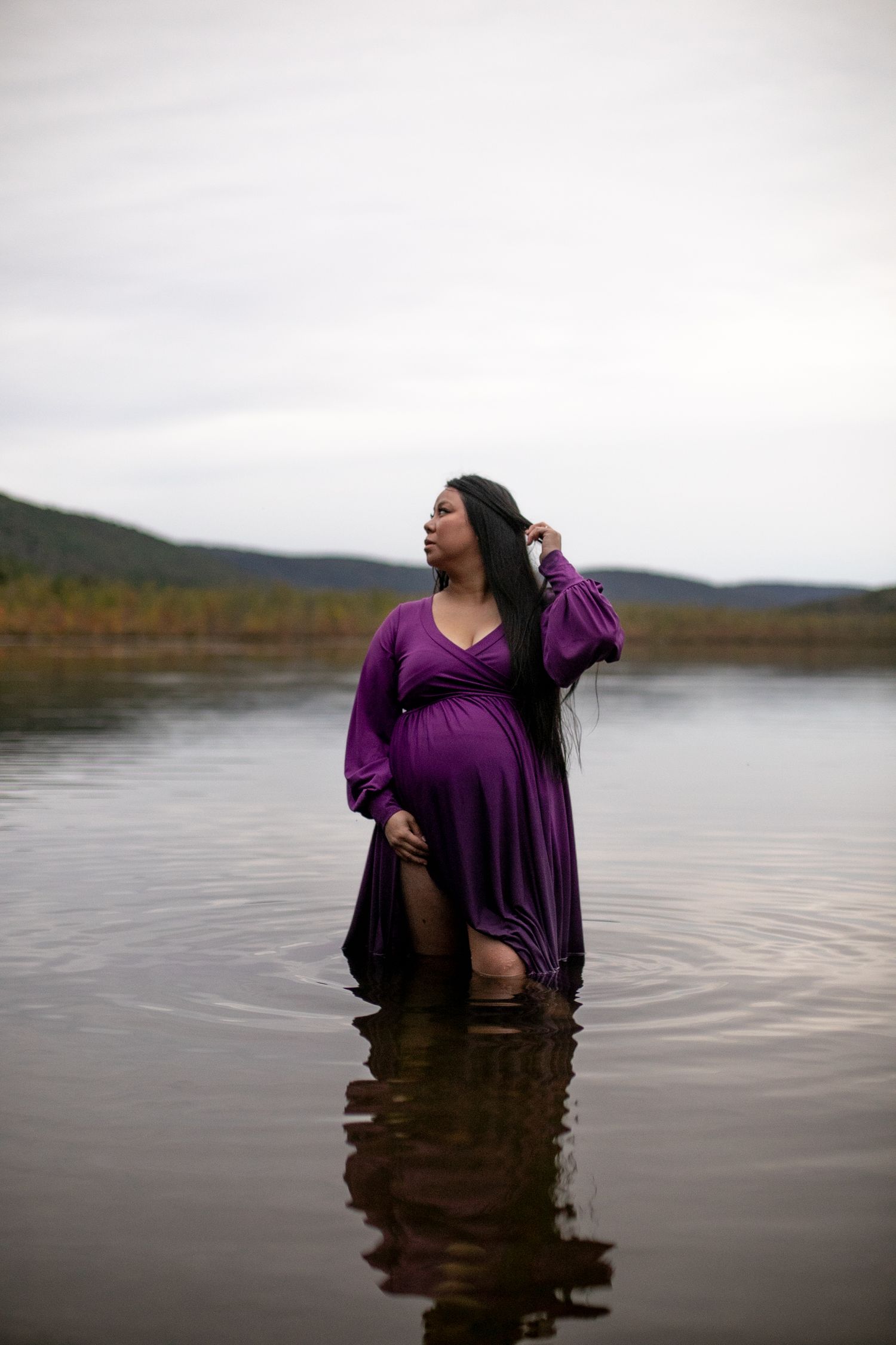 Pregnant woman in purple dress standing in calm lake water, holding hair, with misty hills in background.