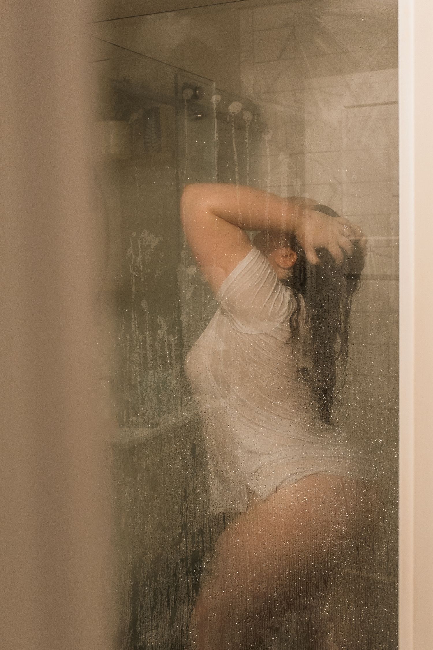 A woman in a wet white bodysuit stands in a steamy glass shower.