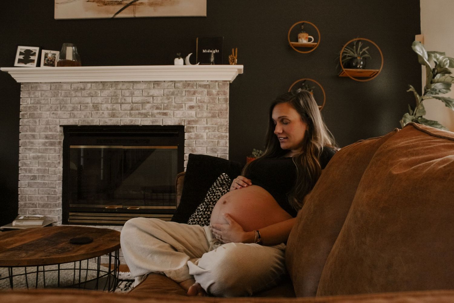 Pregnant woman sitting on floor leaning against couch, cradling baby bump in cozy living room with fireplace.