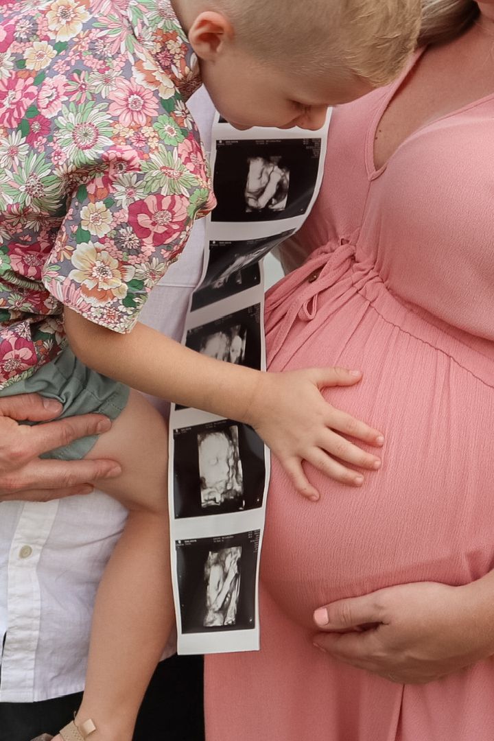 A young child holds ultrasound scans next to a pregnant woman's belly, symbolizing family anticipation of a new baby.