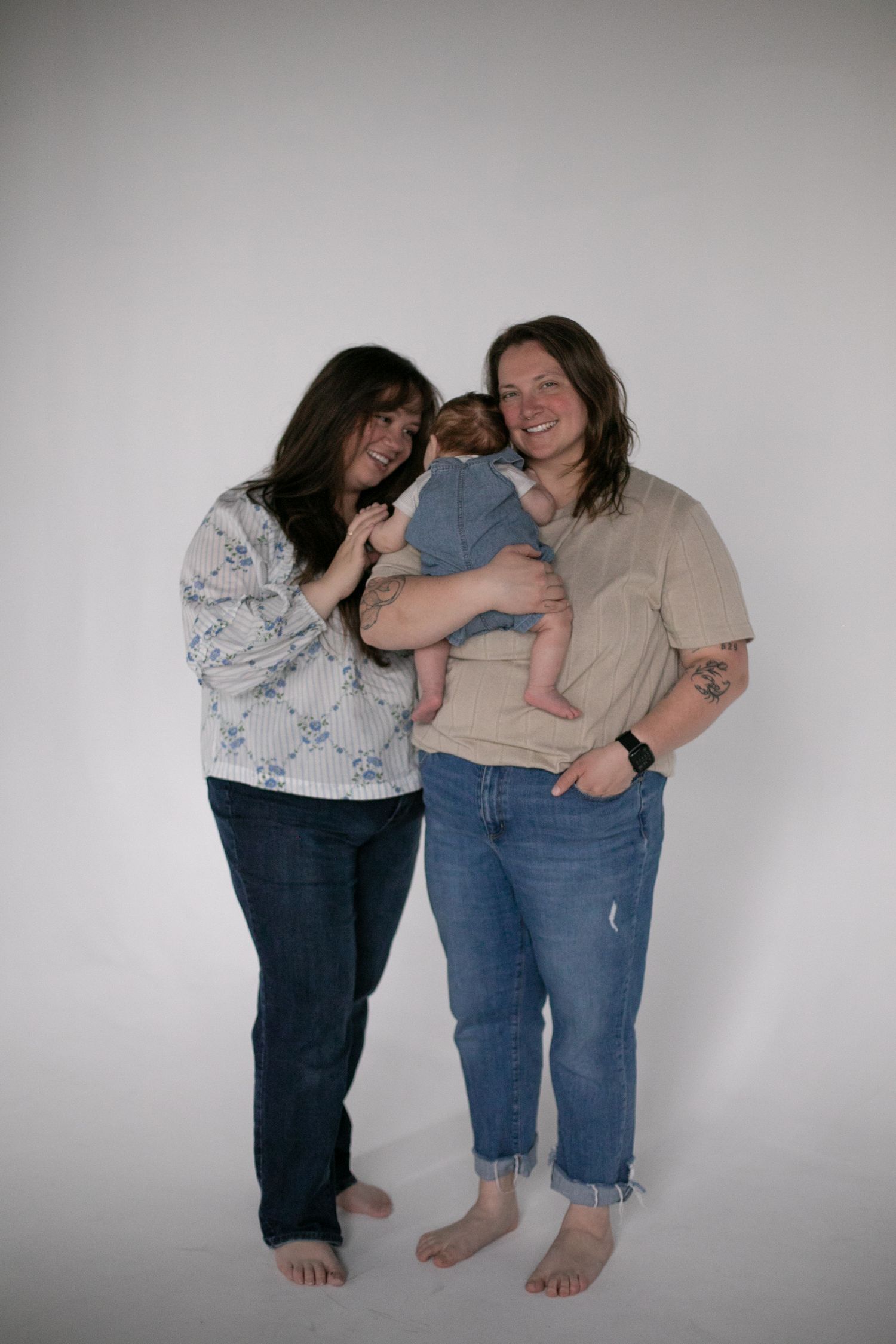 Two barefoot women smiling, holding a newborn baby in a studio portrait setting for motherhood photos by Candid House Studios in Syracuse NY