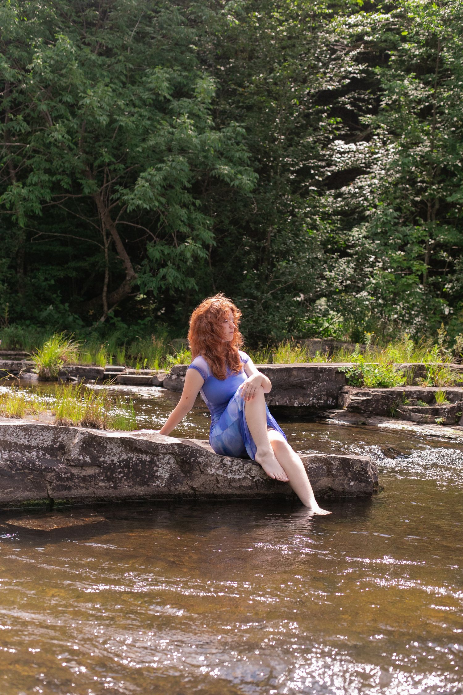 Red-haired woman in blue dress sitting on rocks by a stream, dipping her feet in the water surrounded by lush green trees.