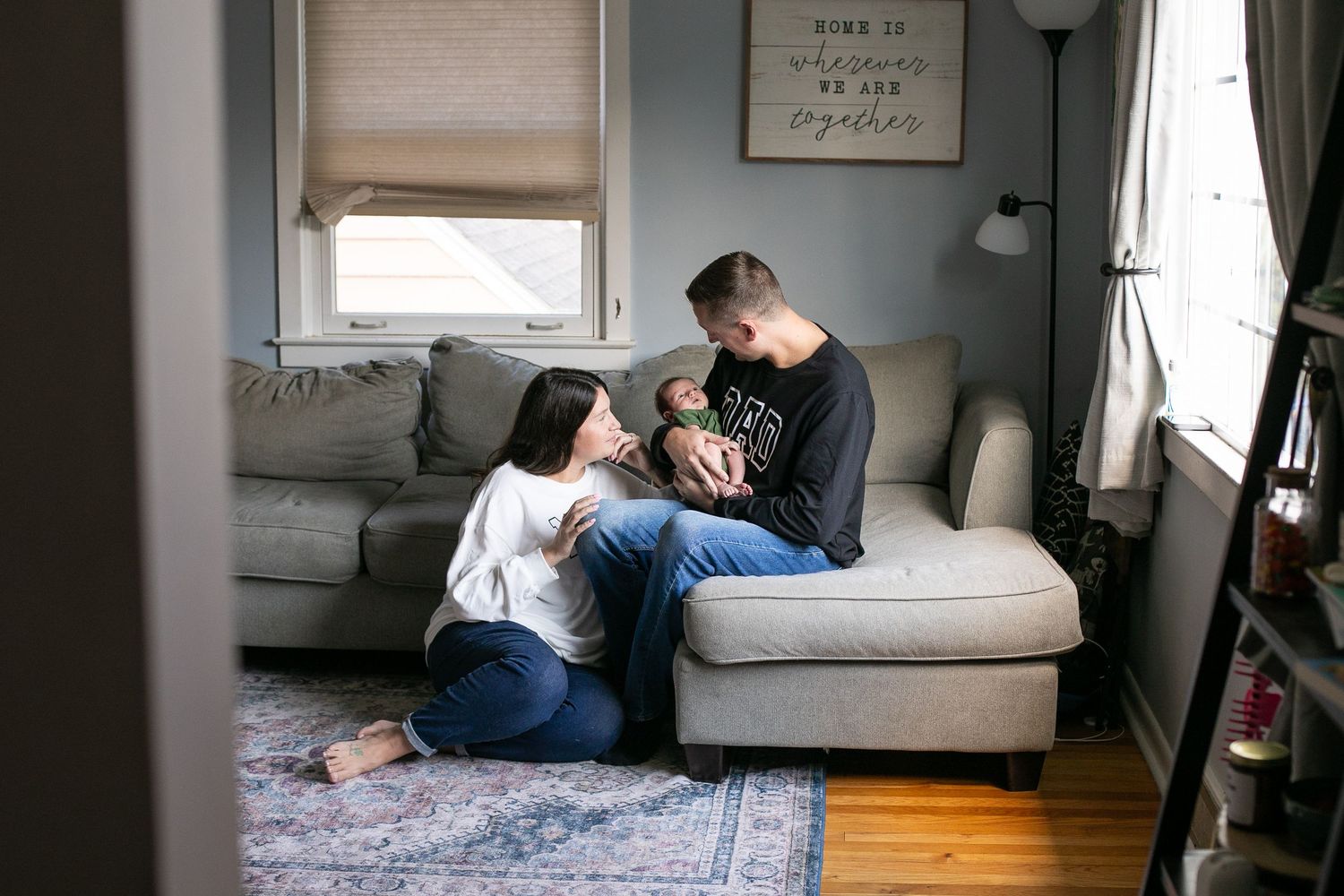 Young family sitting together on living room floor and couch bonding with their newborn baby at home. By Candid House Studios