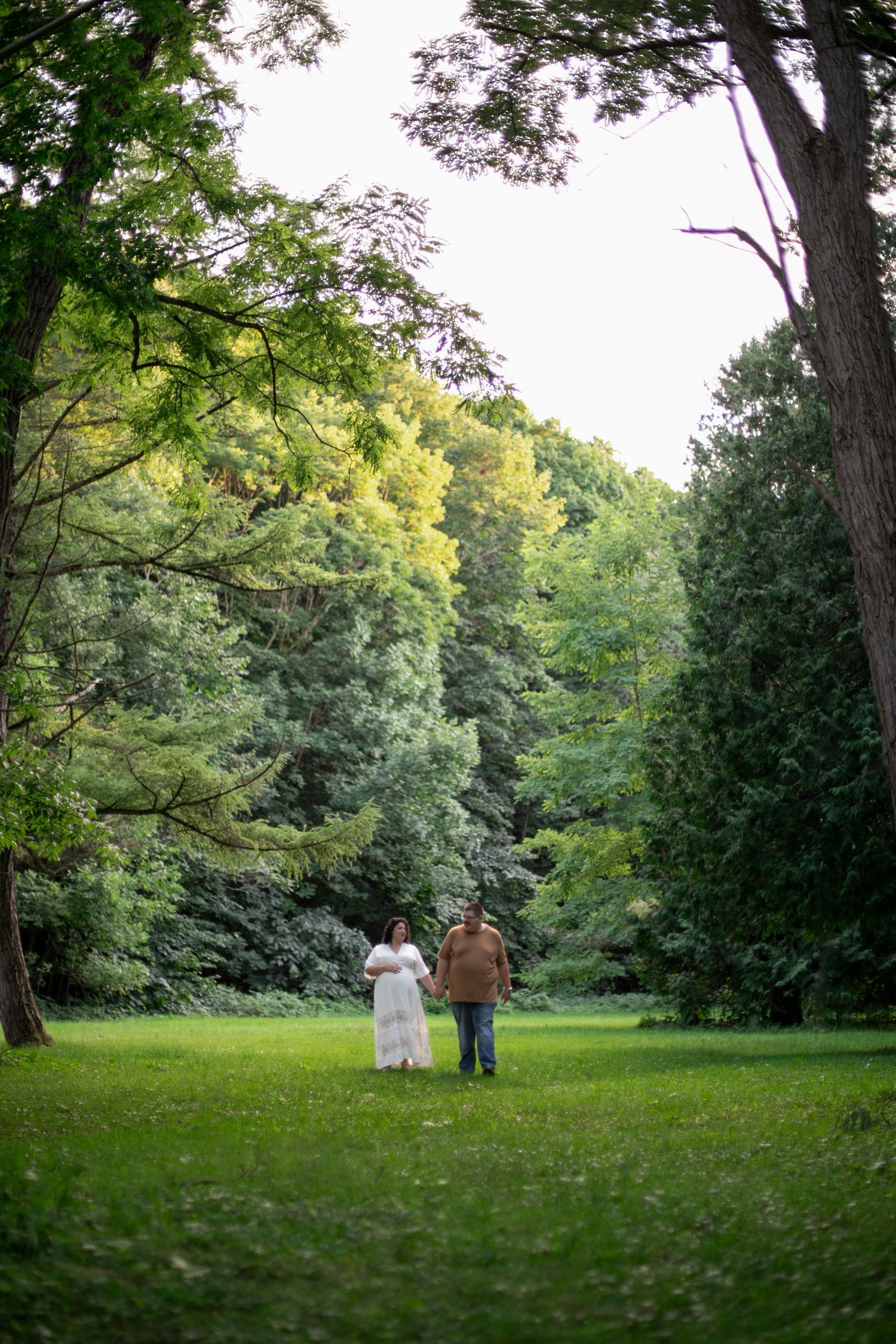 Expecting couple holding hands walking in park, woman in white dress and man in casual attire, during maternity photos by Candid House