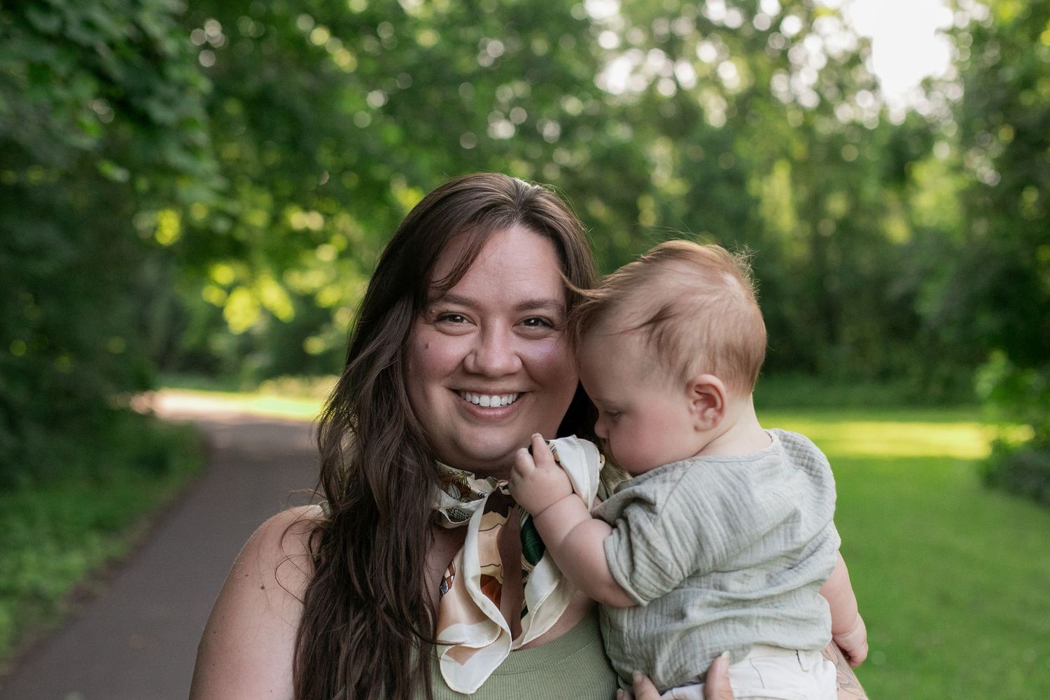 A smiling young woman holds a happy baby outdoors on a lush green park path on a sunny day.
