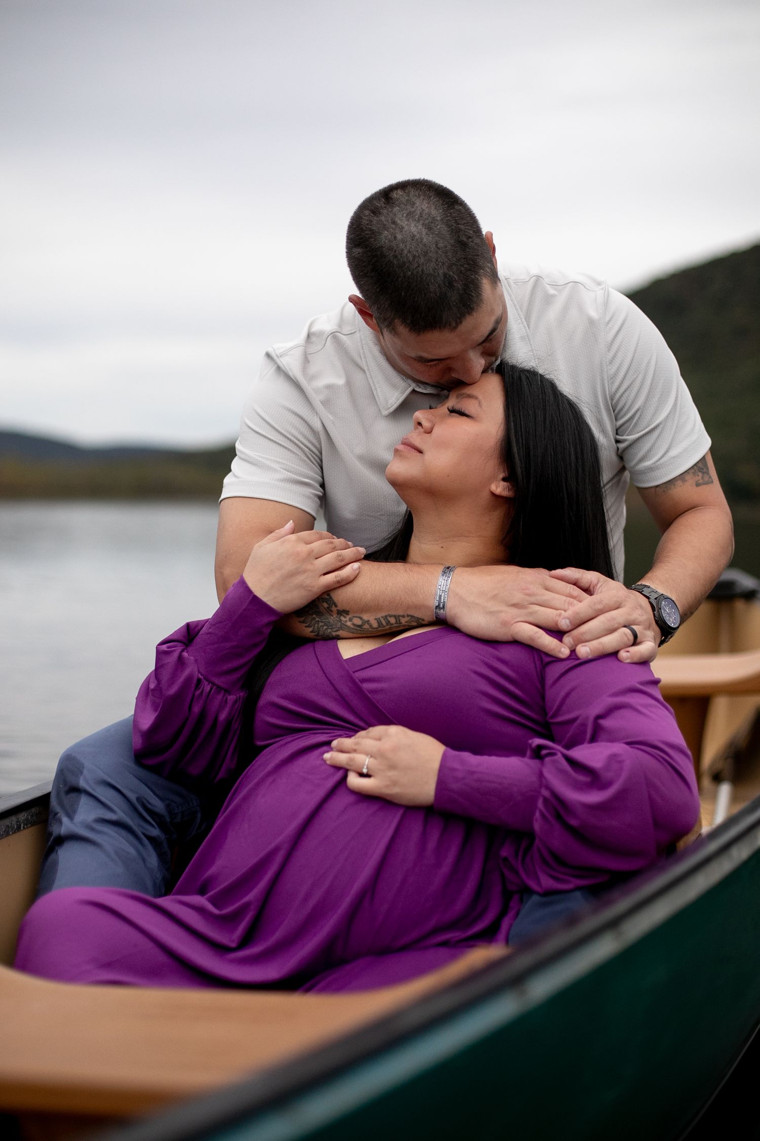 Pregnant woman in purple dress shares a tender kiss with her partner while reclining in a canoe on a serene lake.