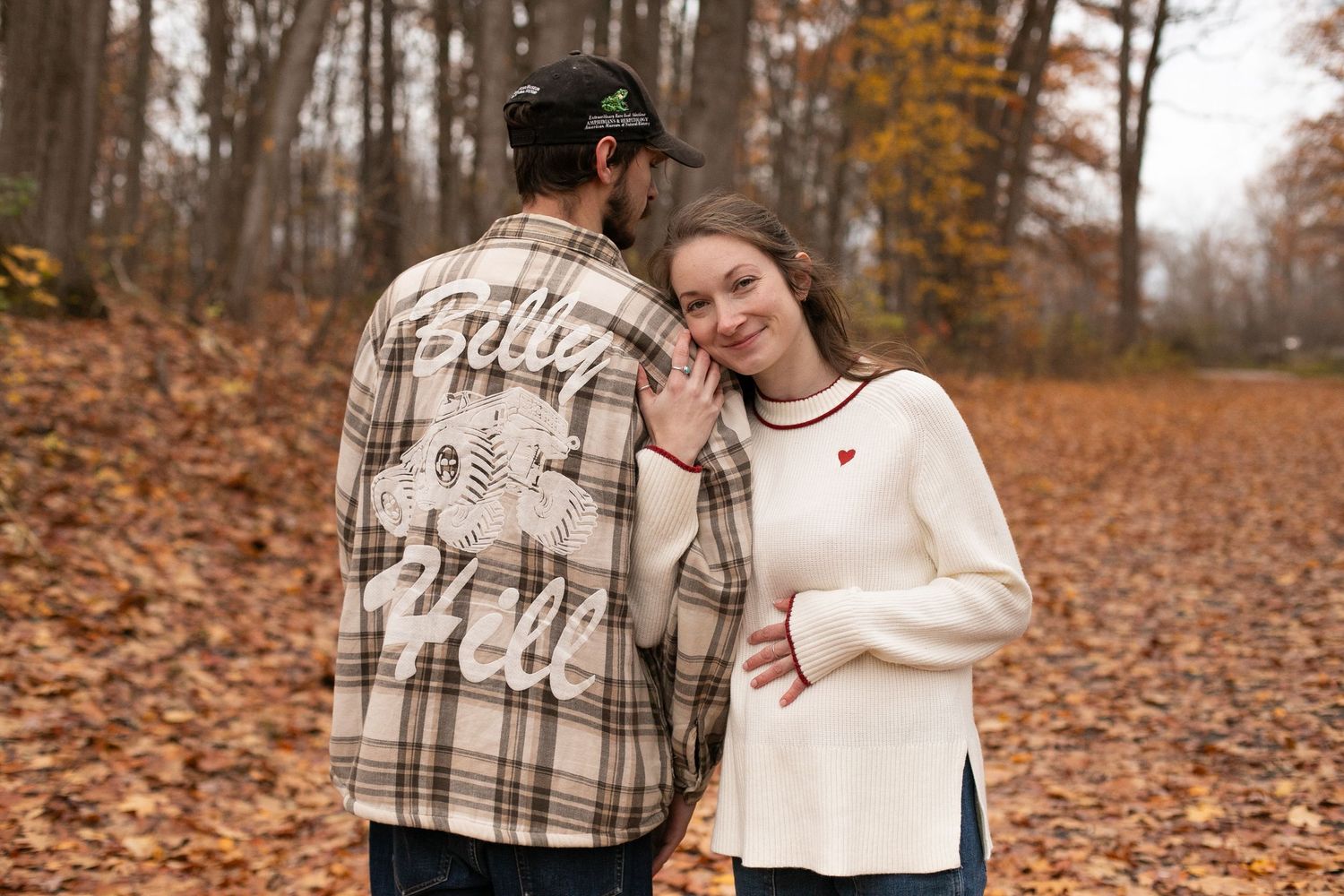 Couple poses in autumn forest, man in plaid flannel shirt, woman in white heart sweater, fall foliage background.