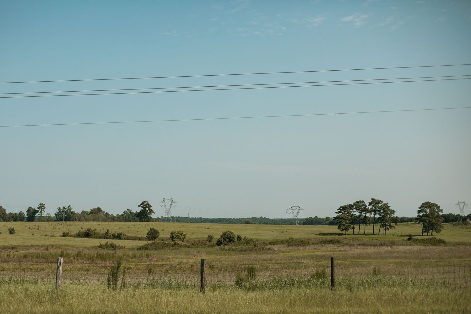 Rural farmland landscape with power lines stretching across flat grassy fields under a clear blue sky.