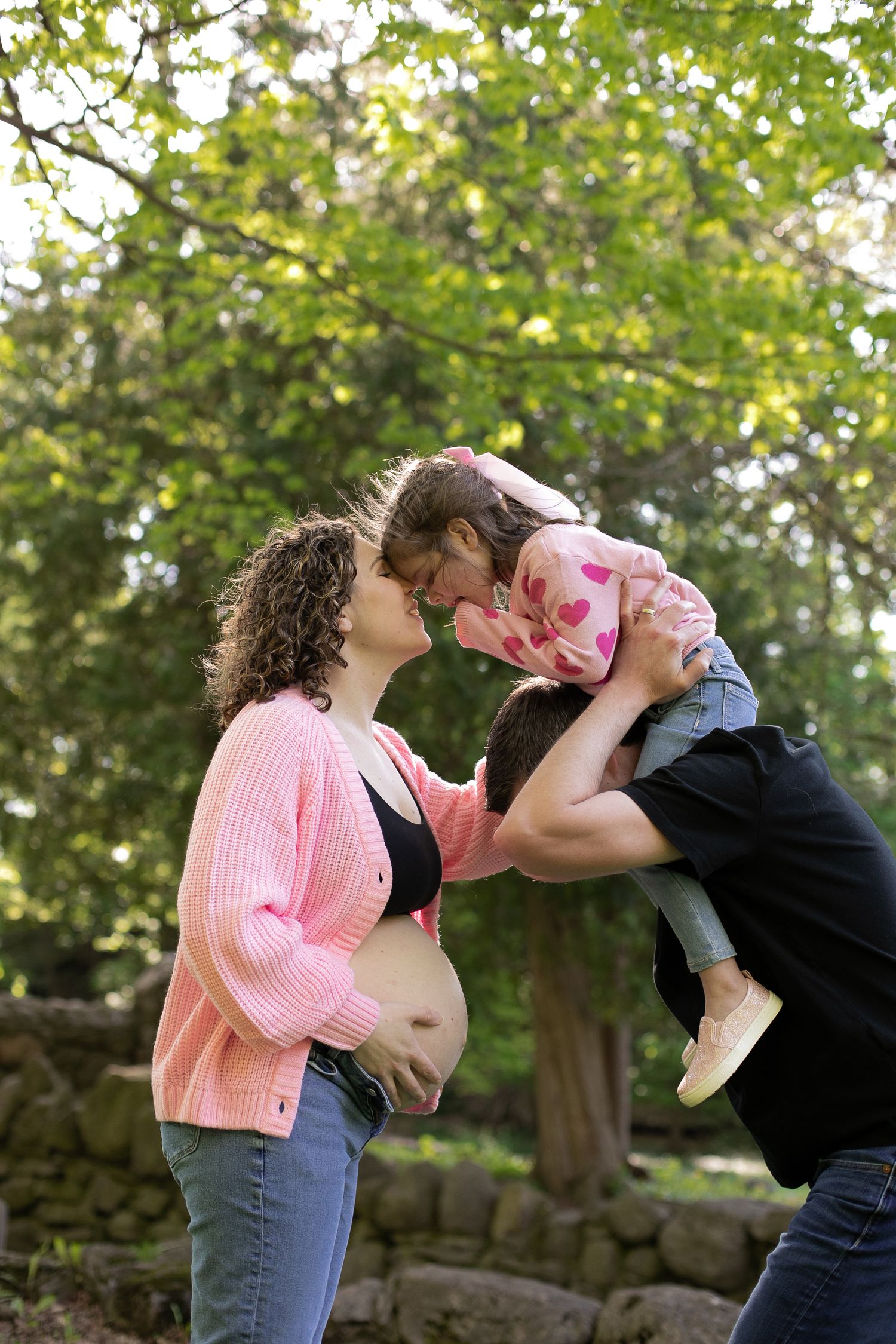 Pregnant woman in pink cardigan shares tender moment with partner lifting young girl in sunny park.