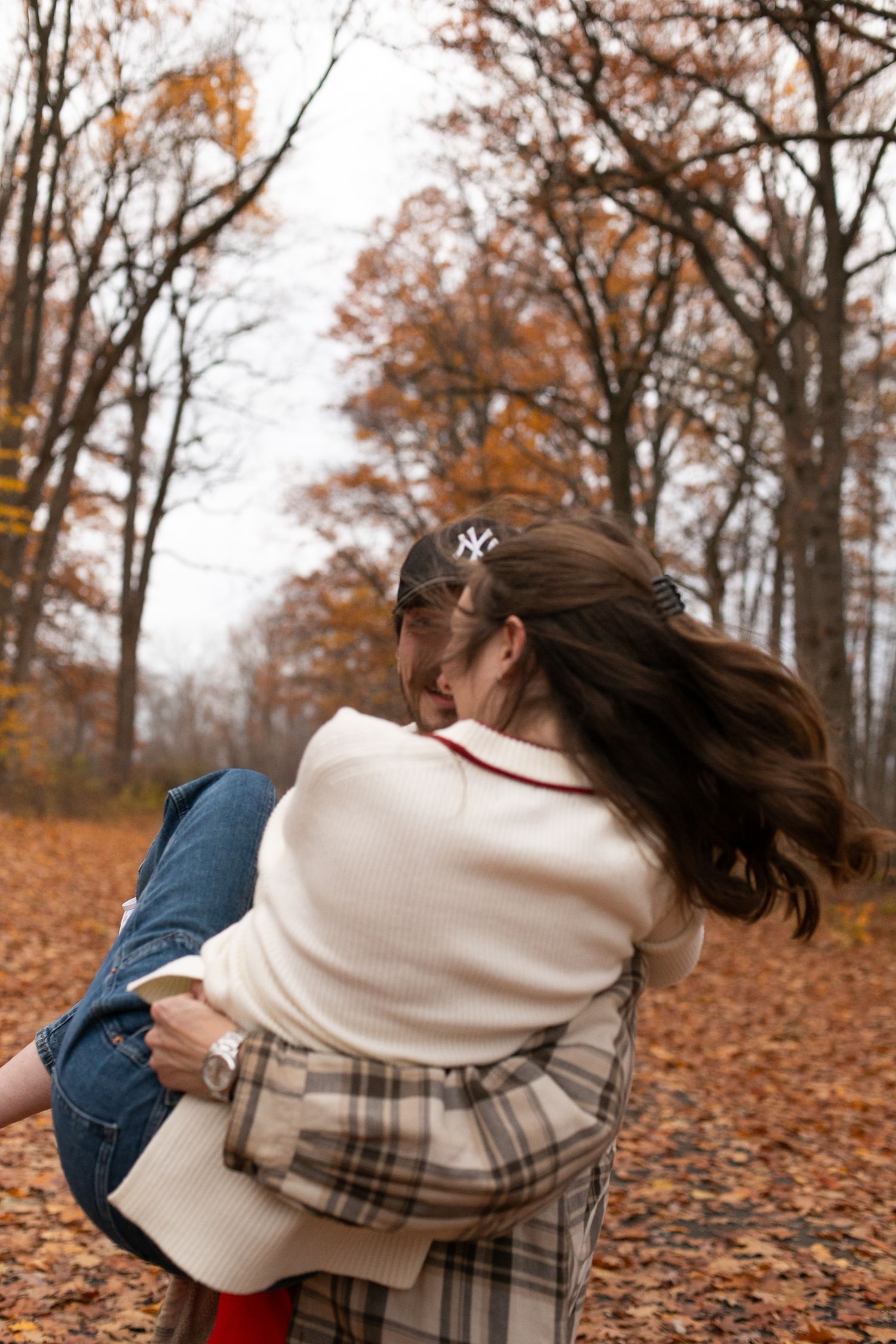 Two women embrace warmly on a leaf-covered autumn path surrounded by colorful fall foliage trees.
