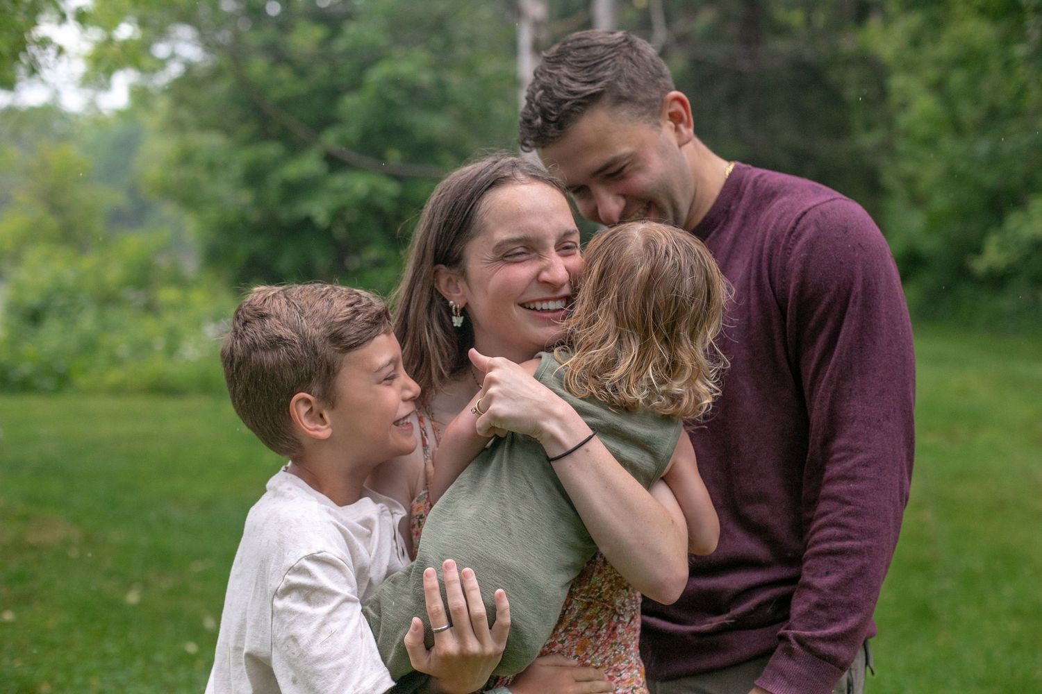 Happy family of four sharing warm embrace outdoors in green park, parents hugging children lovingly. By Candid House Studios, Syracuse, NY