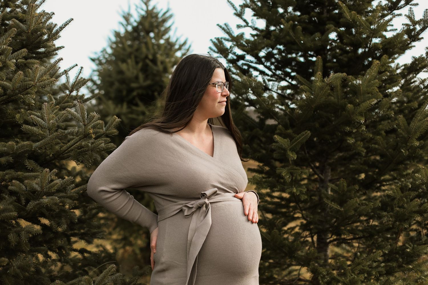 Pregnant woman in taupe wrap dress poses among evergreen trees at a Christmas tree farm maternity photoshoot.
