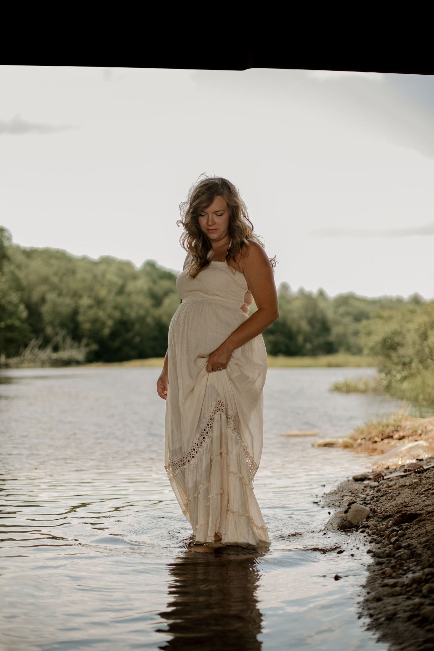 Pregnant woman in white boho dress standing in shallow river water surrounded by lush green trees.