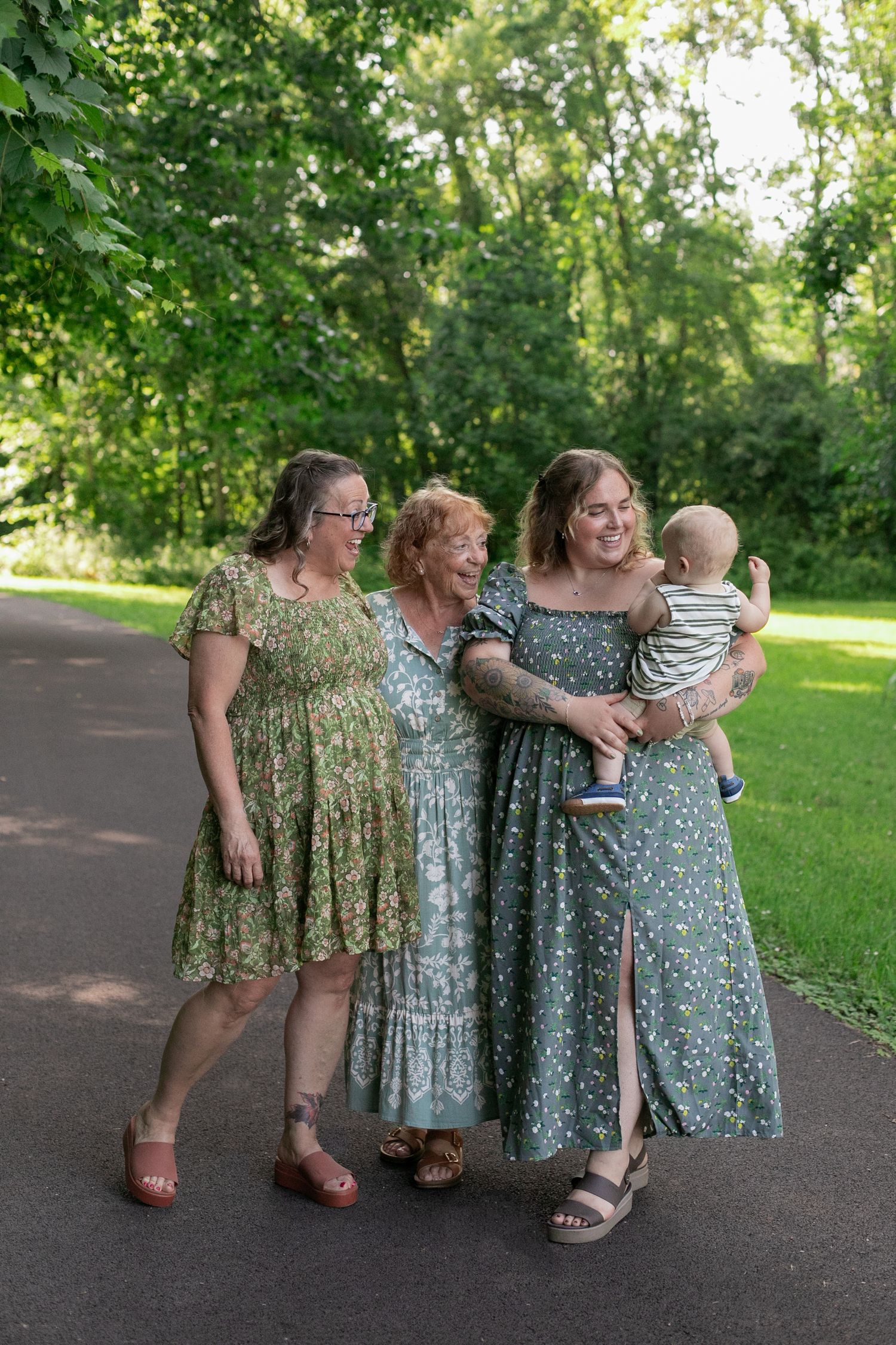 Three women in matching green floral dresses walk outdoors, one holding a baby, for their Legacy generations photos by Candid House Studios.