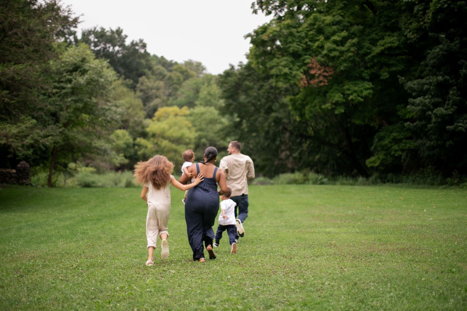 Family walking together across lush green park lawn surrounded by tall trees during family photoshoot by Candid House Studios, Syracuse, NY.