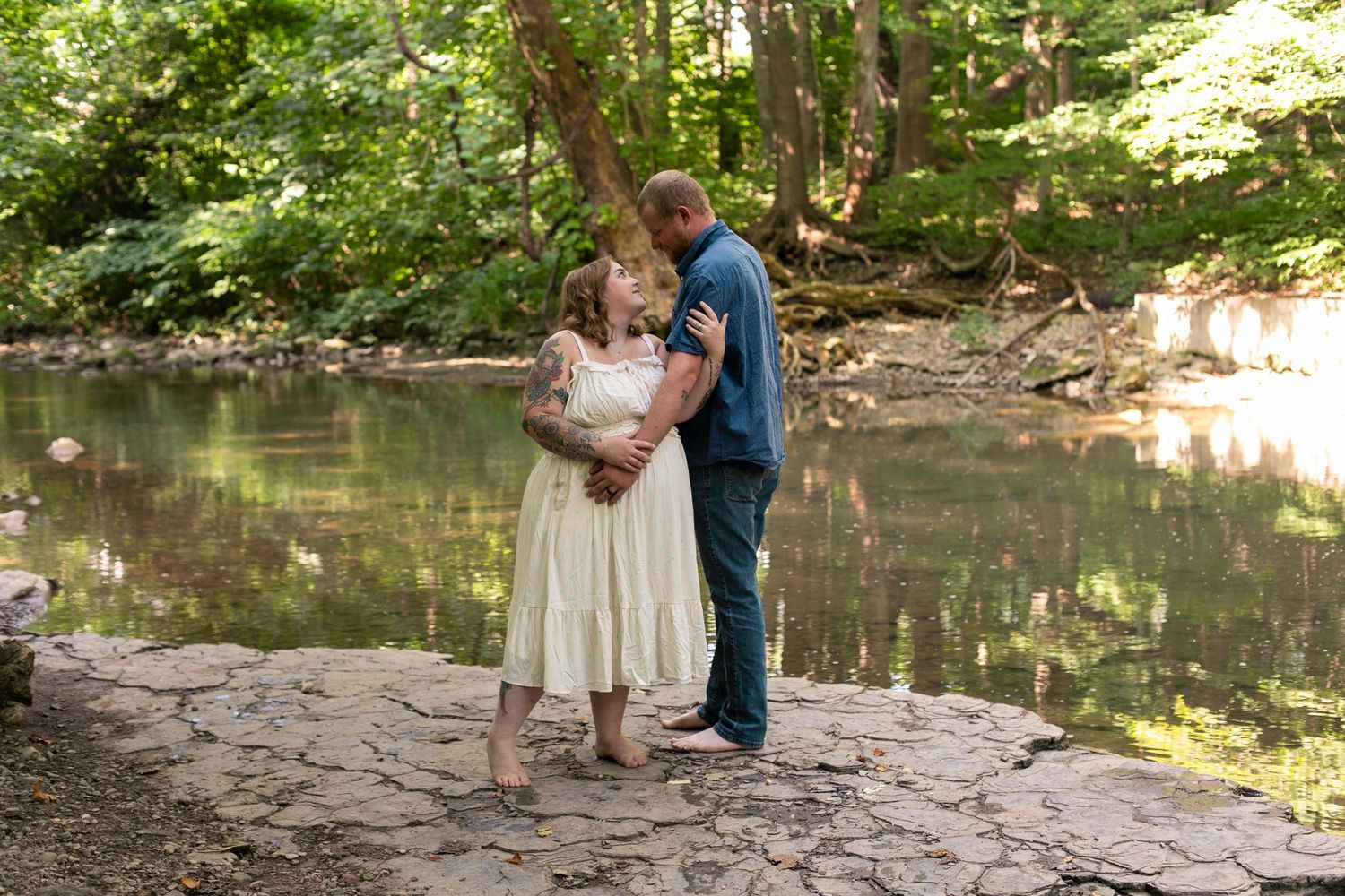 Couple embracing tenderly by a serene forest creek on flat rocks, woman in white dress, man in teal shirt, lush green trees.