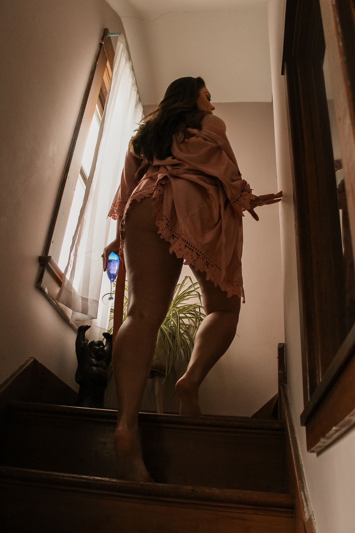 A woman in a flowing pink robe stands barefoot on wooden stairs near a sunlit window.