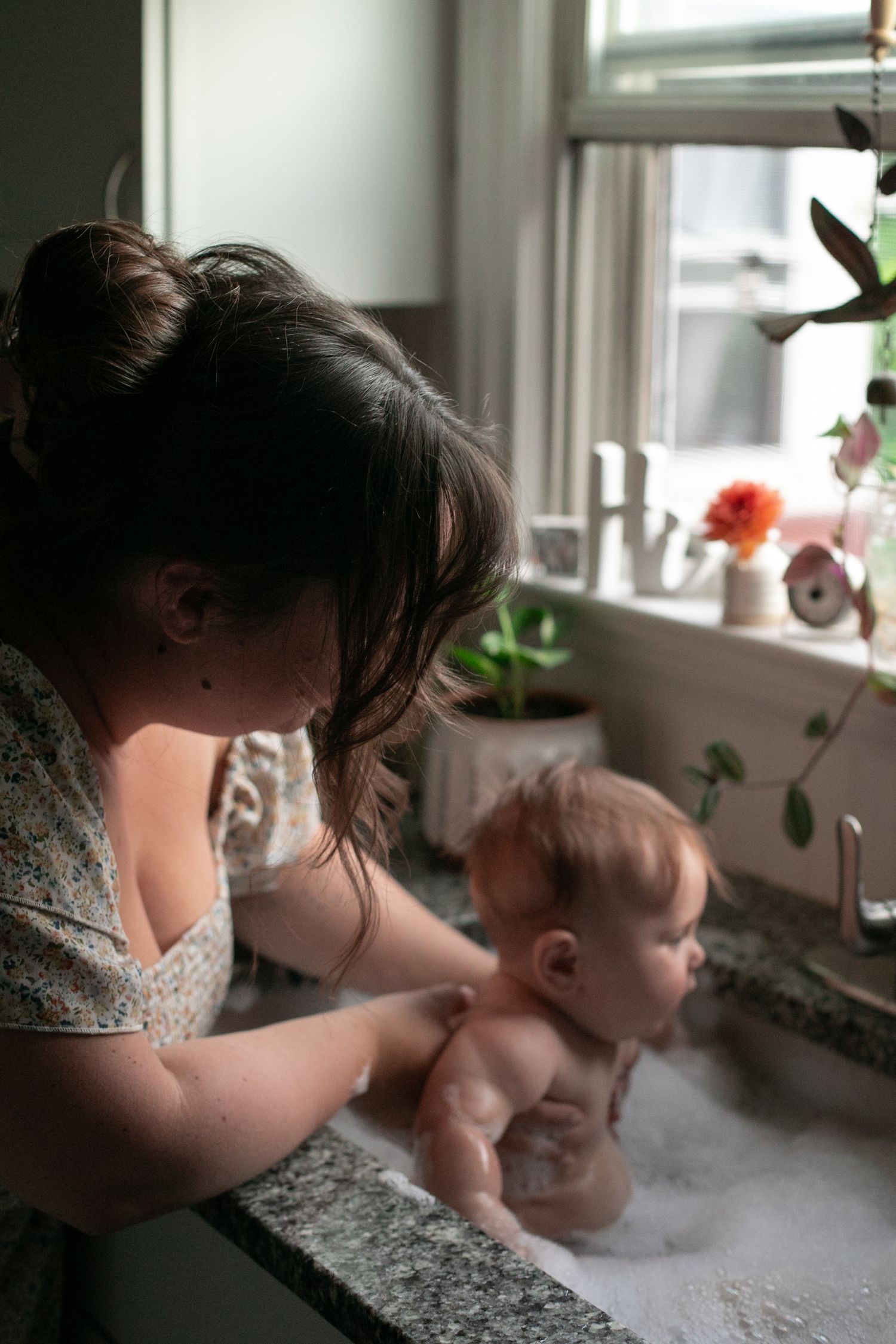 Mother gently bathing infant in kitchen sink filled with bubbles near sunlit window with plants. By Candid House Studios, Syracuse, NY