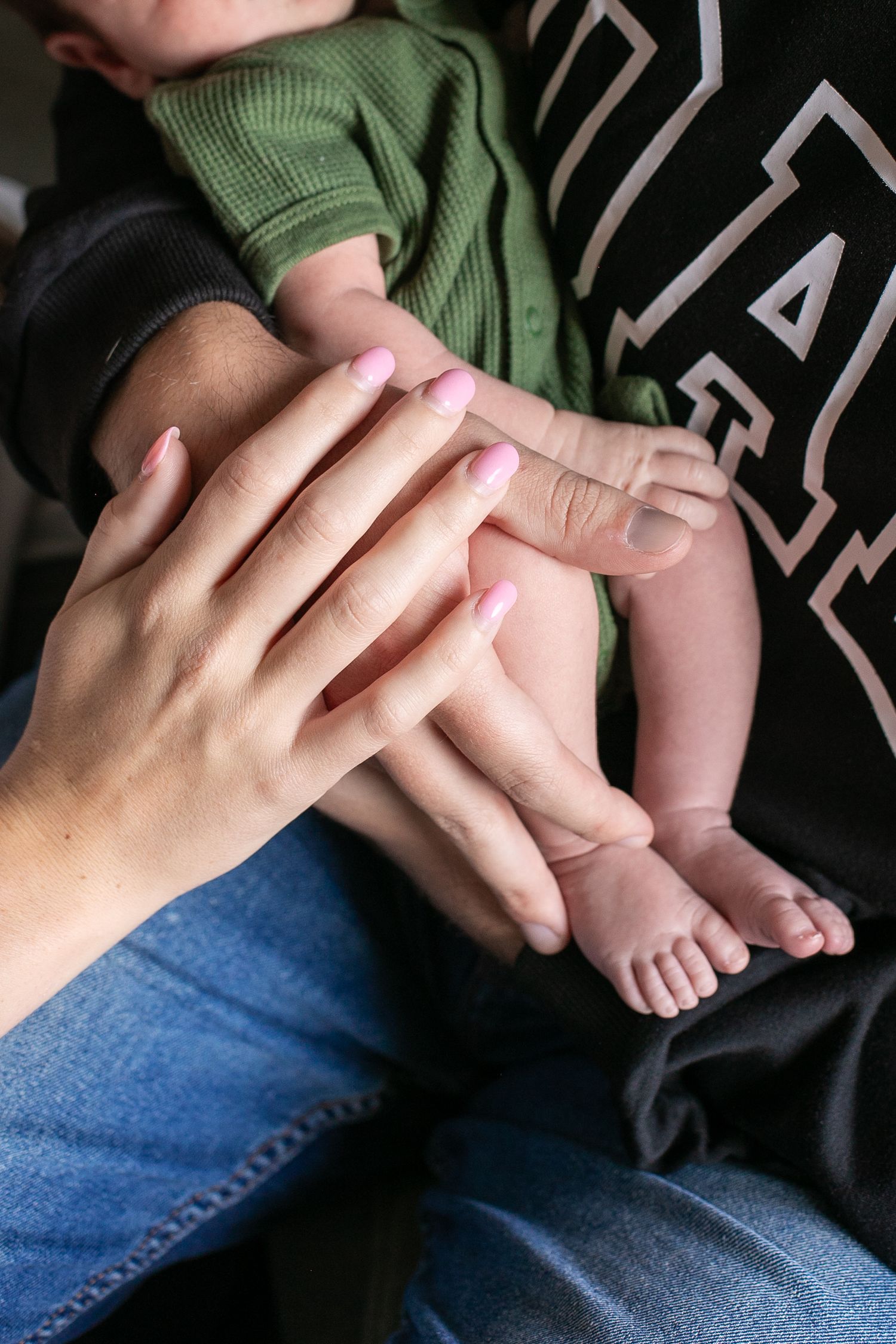 Close-up of mother and father's hands gently holding newborn baby's tiny feet and legs together. By Candid House Studios, Syracuse NY