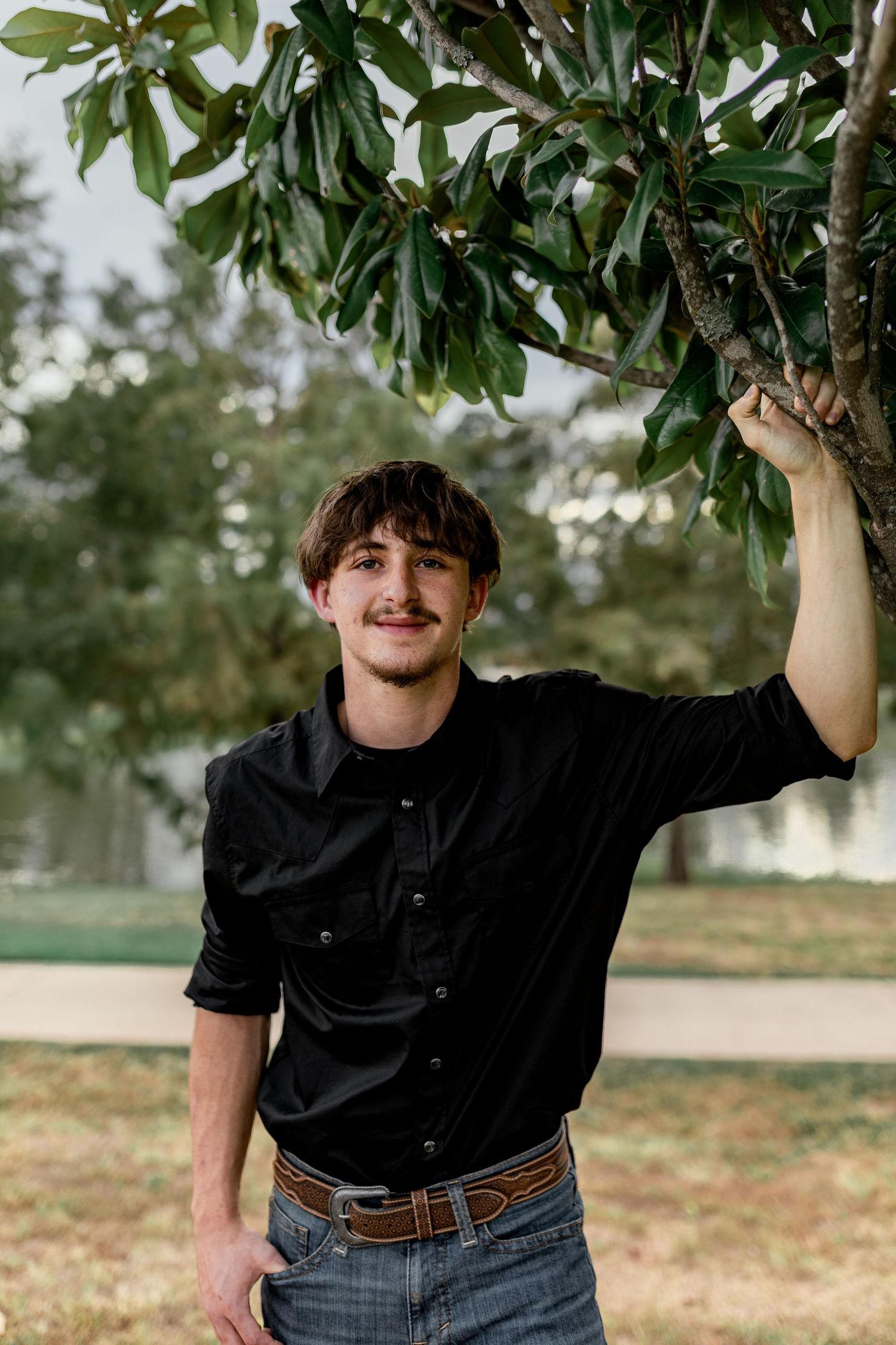 Smiling young man in black shirt holding magnolia tree branch, standing in a green outdoor park.