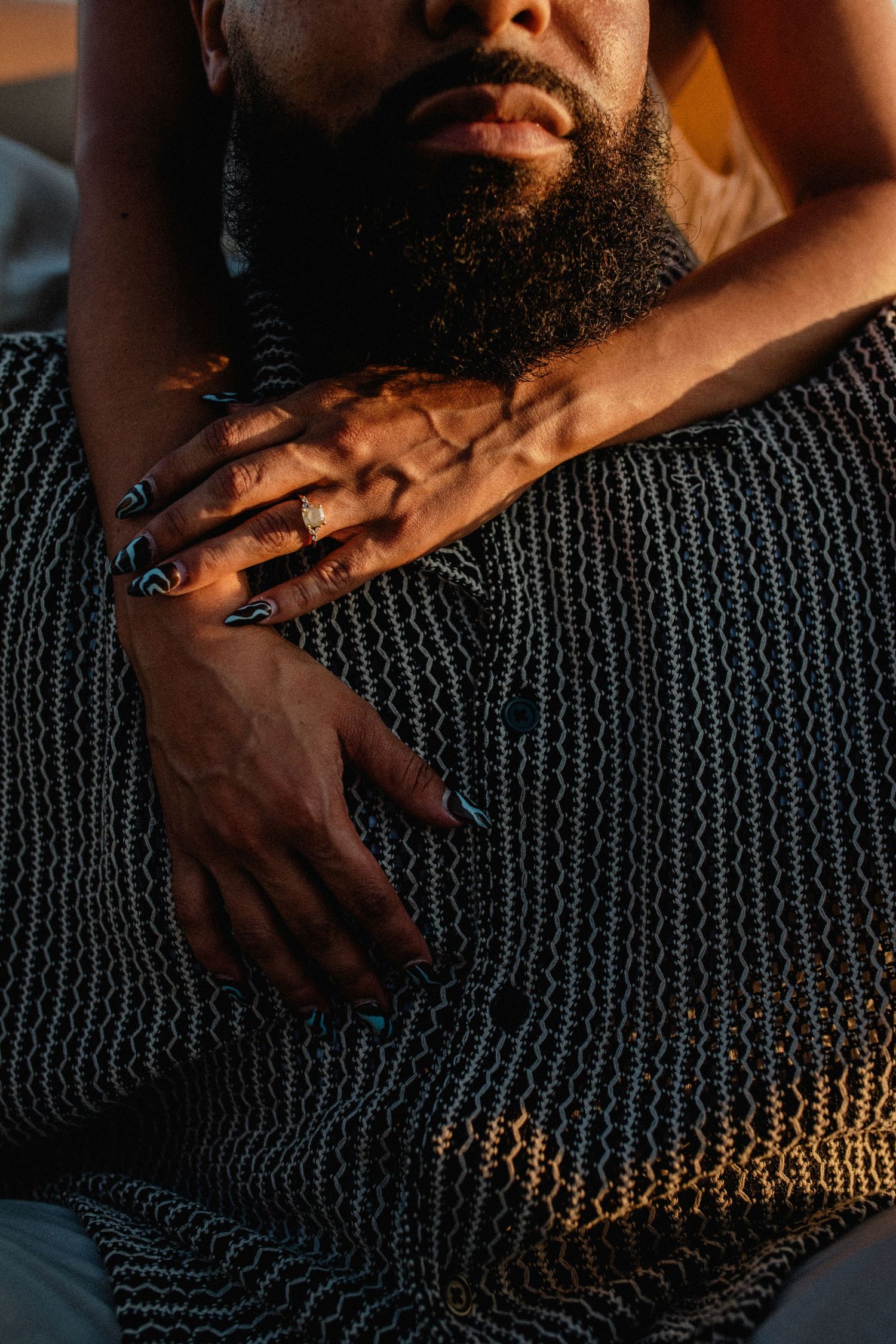 Close-up of womans hands with dark nails resting on bearded mans chest in warm light.