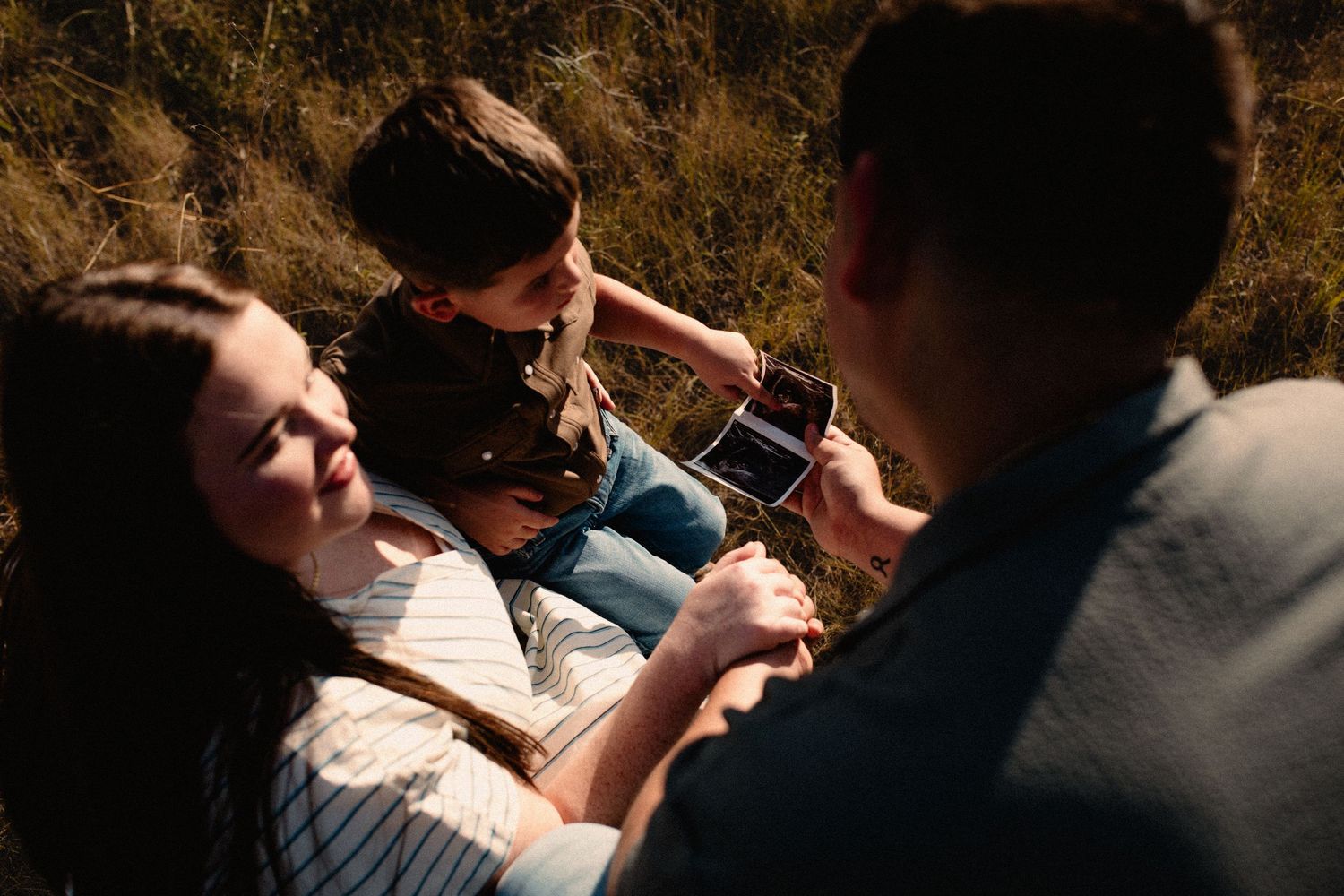 Family sitting in grass as young boy shows parents an ultrasound photo during golden hour.