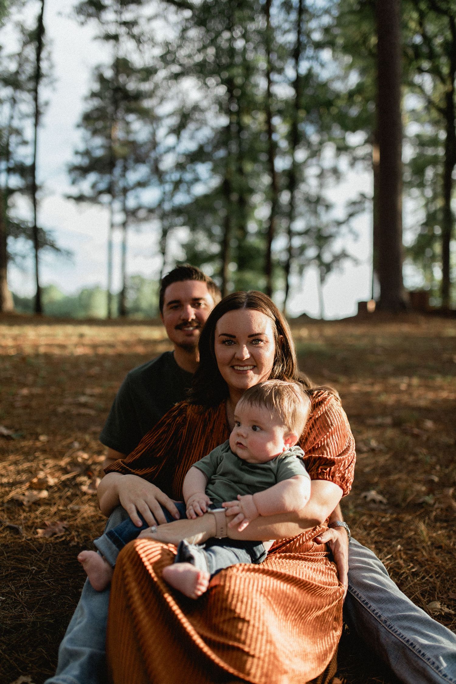 A smiling family sits together on pine needle ground, with tall trees and sunlight behind them.