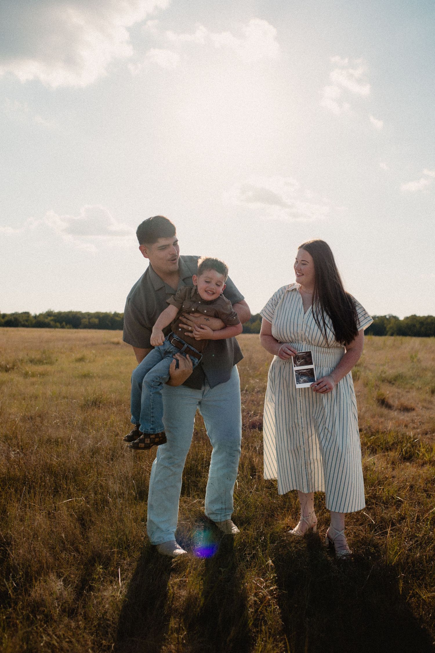 Happy family pregnancy announcement in field, father holding son as mother shows ultrasound photos.