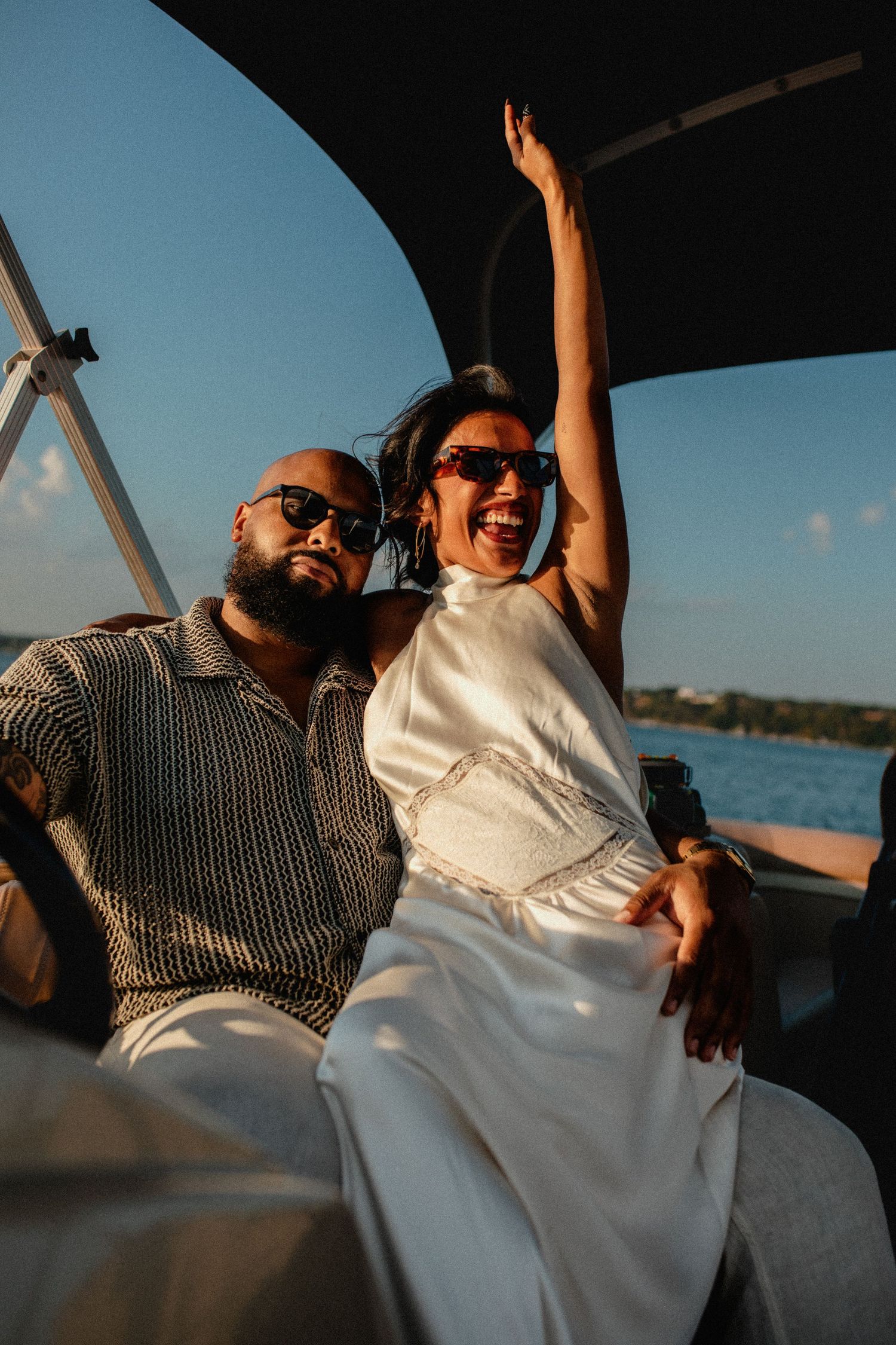 Excited couple on a boat at sunset, woman raising arm in celebration wearing sunglasses and white dress.