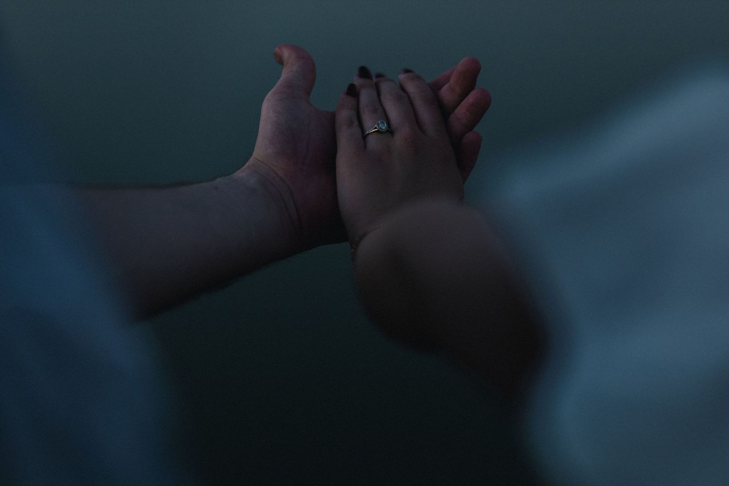 Woman's hand with ring gently touching fiancee's hand in soft dim light, symbolizing love and engagement.