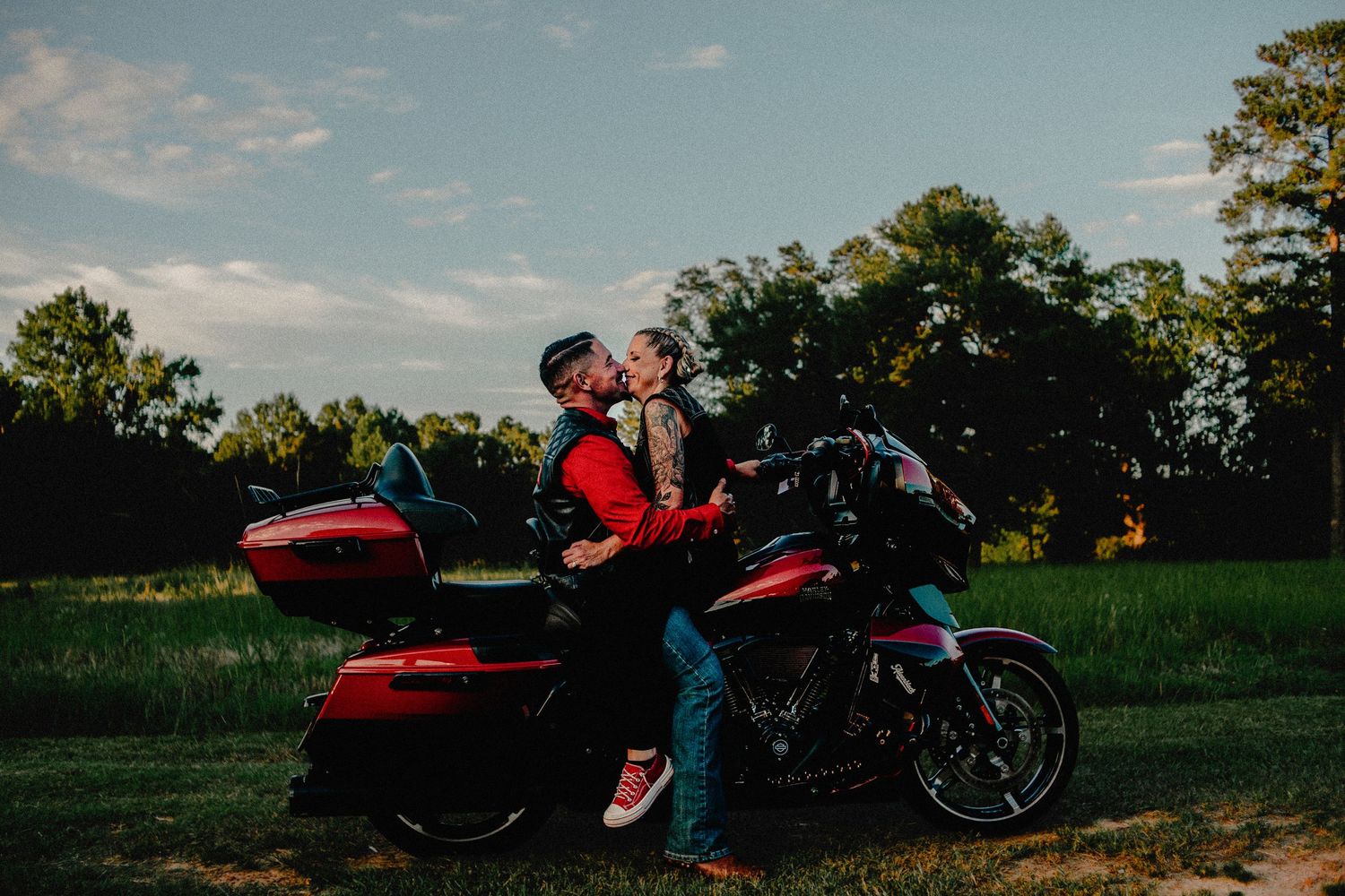 Couple sharing a romantic kiss leaning against a red Harley-Davidson motorcycle in a scenic green field at sunset.