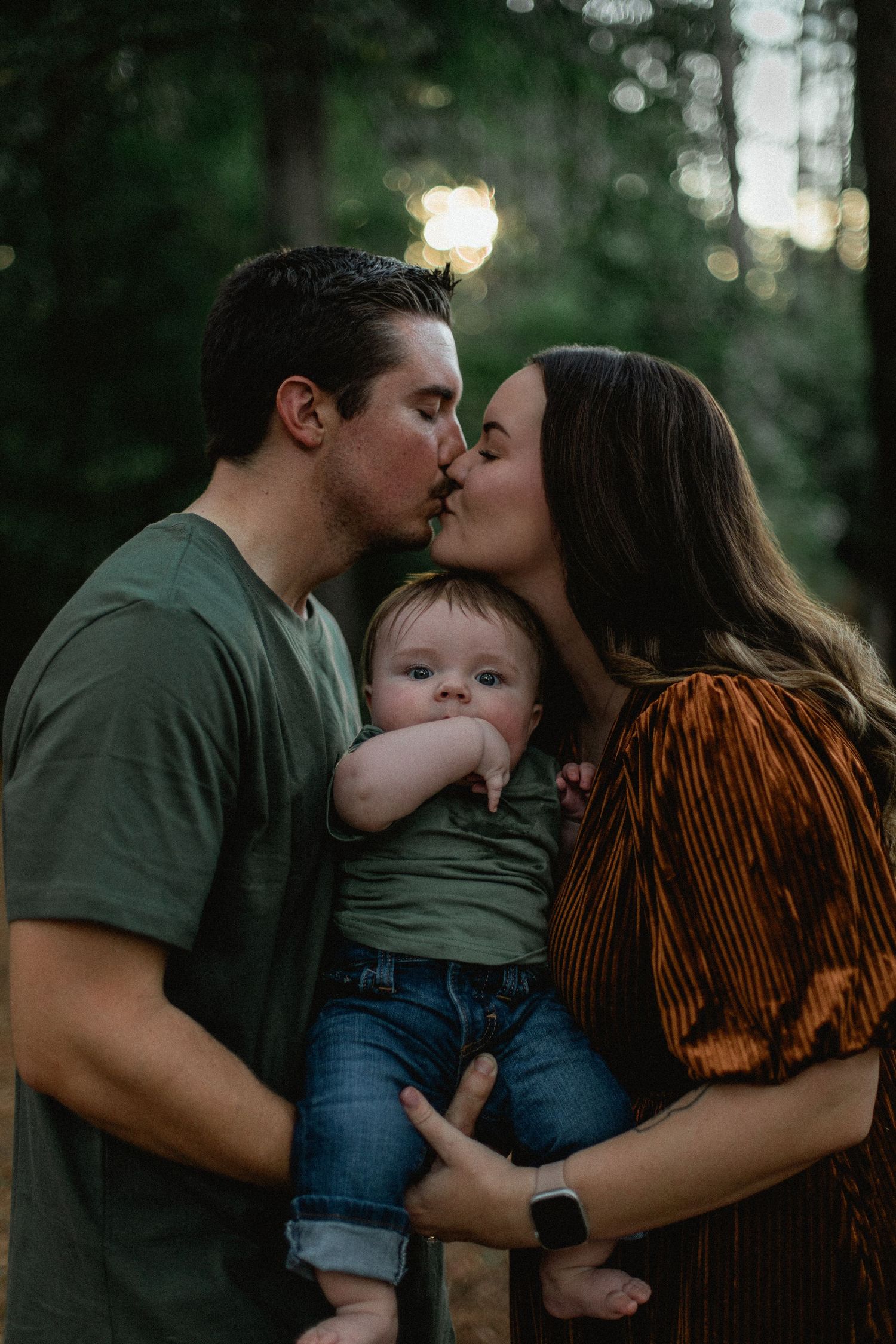 Happy couple kissing while holding their baby boy in a sunlit forest setting.