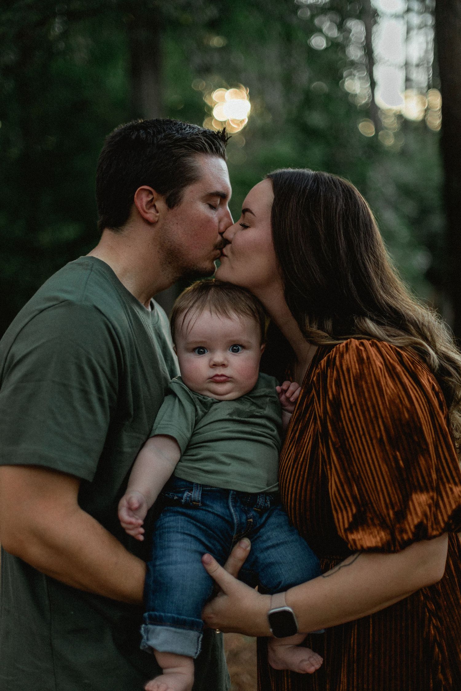 Couple sharing a kiss while holding their baby between them in a lush forest.