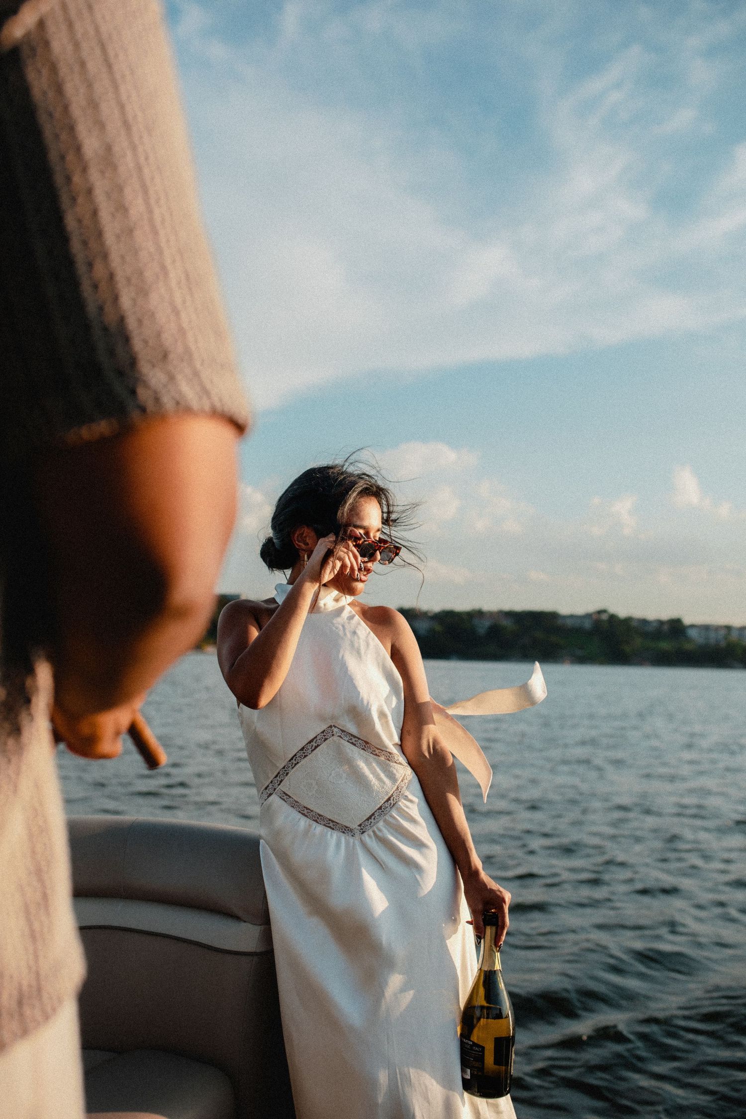 Woman in white halter dress holding champagne bottle on a boat, wind blowing her hair on a sunny day.