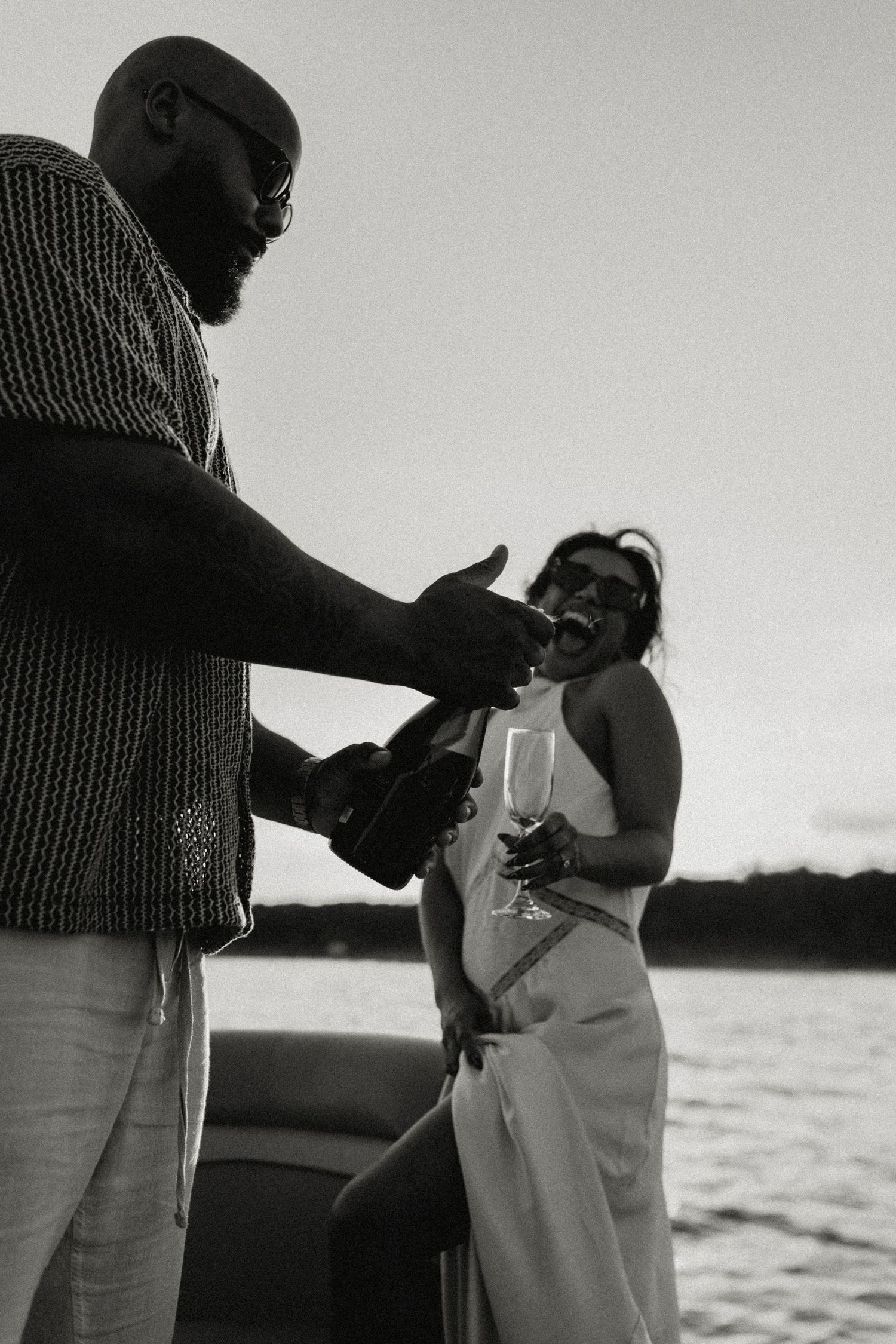 Black and white photo of couple opening champagne on a boat at sunset.
